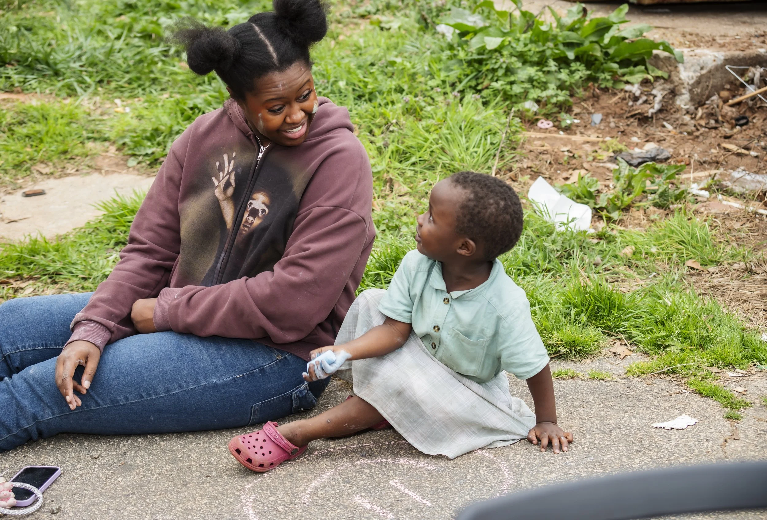 A woman with dark hair in buns, wearing a brown hoodie with images on the front, is sitting on the ground near a young boy. The boy, wearing a light green shirt, plaid skirt, and pink Crocs, is kneeling on the pavement and holding a piece of chalk. T
