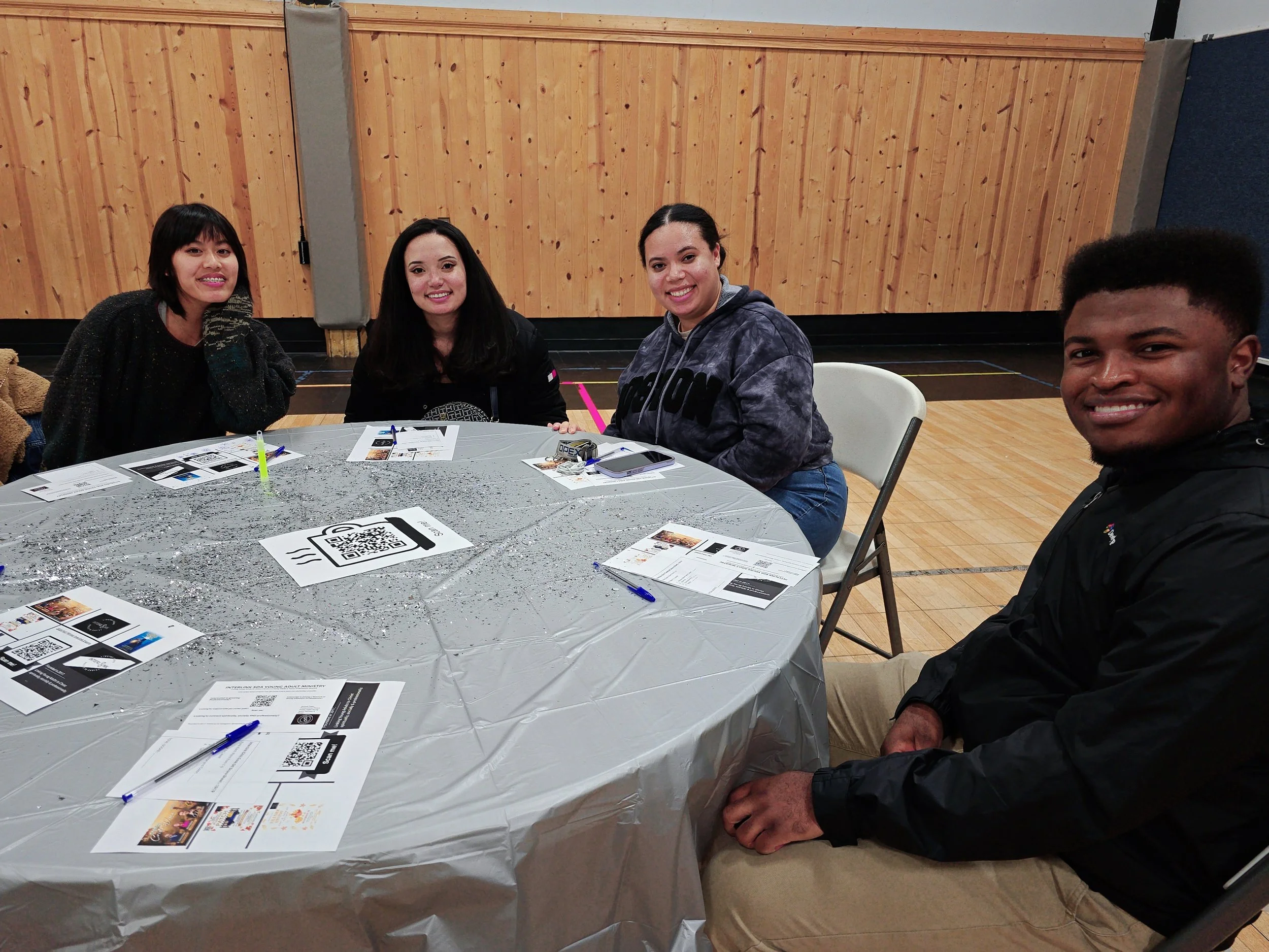 Four young adults sit at a round table covered with a plastic tablecloth and printed papers, smiling at the camera in a room with wooden walls and a hardwood floor.