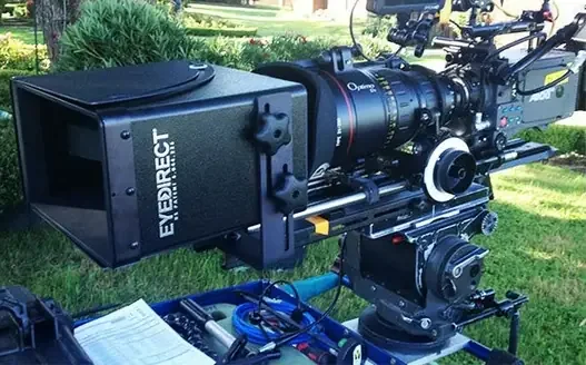EyeDirect - Large professional video camera mounted outdoors on a tripod with greenery in the background.