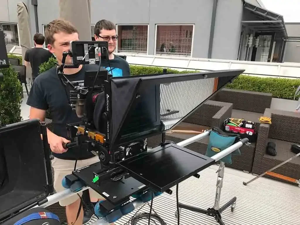Two men operating a large camera on a steadycam rig on a rooftop patio with outdoor furniture and items in background.