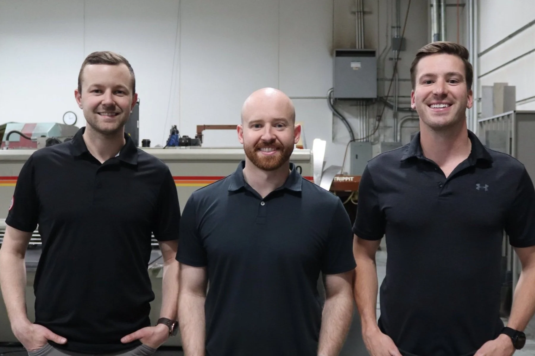Three men in black polo shirts standing in an industrial setting, smiling.
