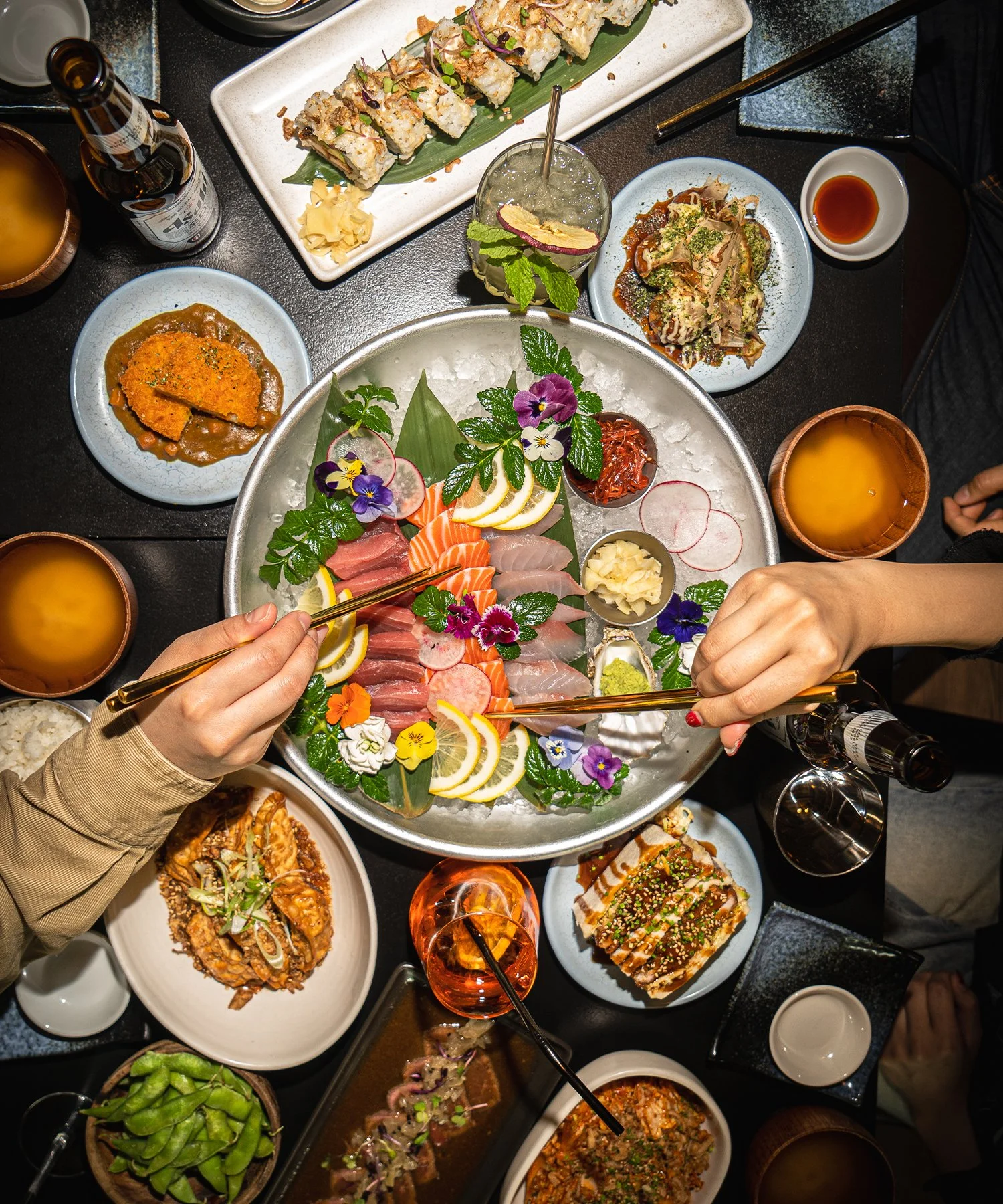 A large platter of assorted sashimi with colorful edible flowers, lemon slices, and garnishes, surrounded by various Japanese dishes including sushi, tempura, and drinks on a black table.