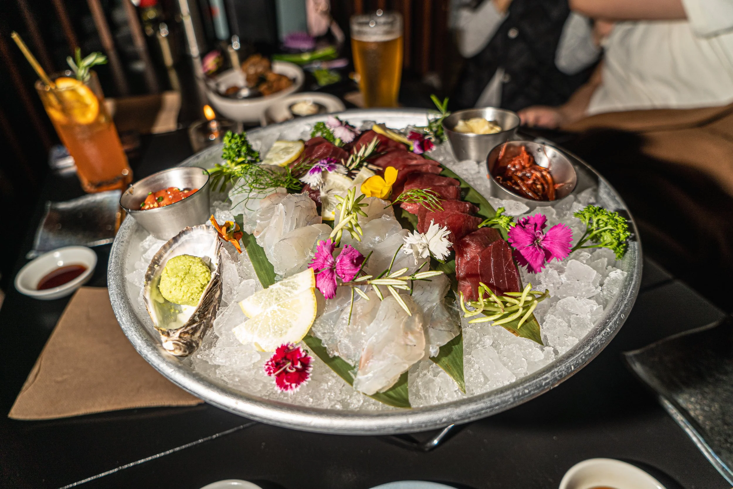 A large round silver tray filled with ice and topped with assorted sashimi including slices of white fish and red tuna, garnished with flowers, lemon wedges, and herbs. Several small bowls with dipping sauces and condiments surround the tray, with dr