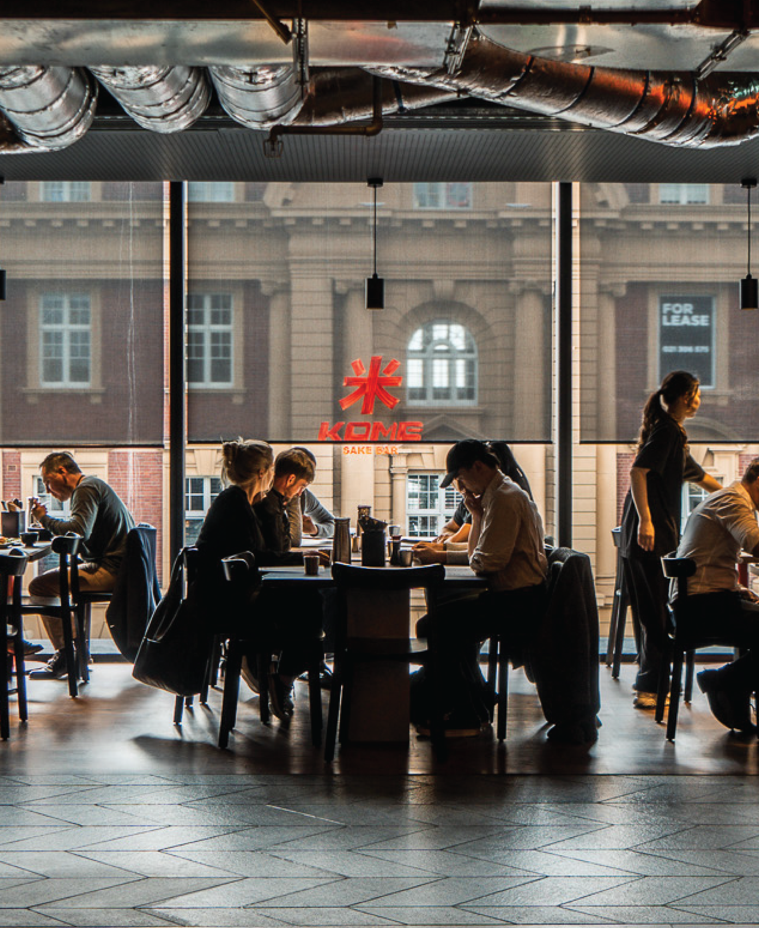 People sitting and working at tables inside a cafe with large windows, city buildings visible outside.