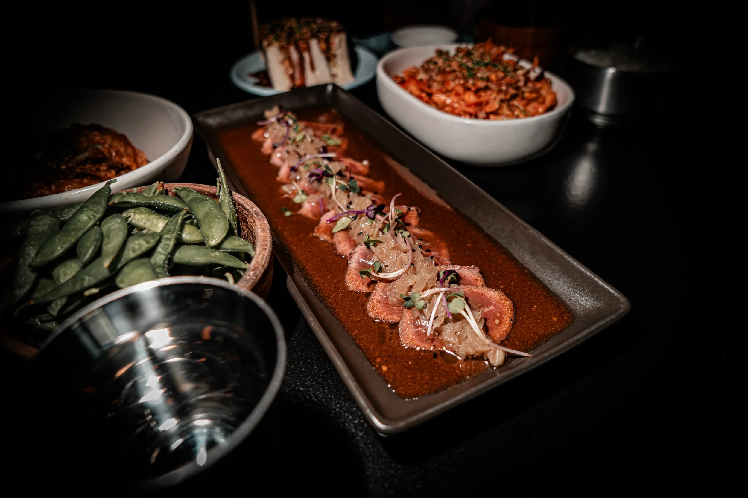 A long rectangular plate with slices of beef topped with onions and microgreens in a dark sauce. Around the plate, there are bowls of edamame, a layered dessert, a spicy tofu dish, and a piece of sliced cake, all set on a dark table.
