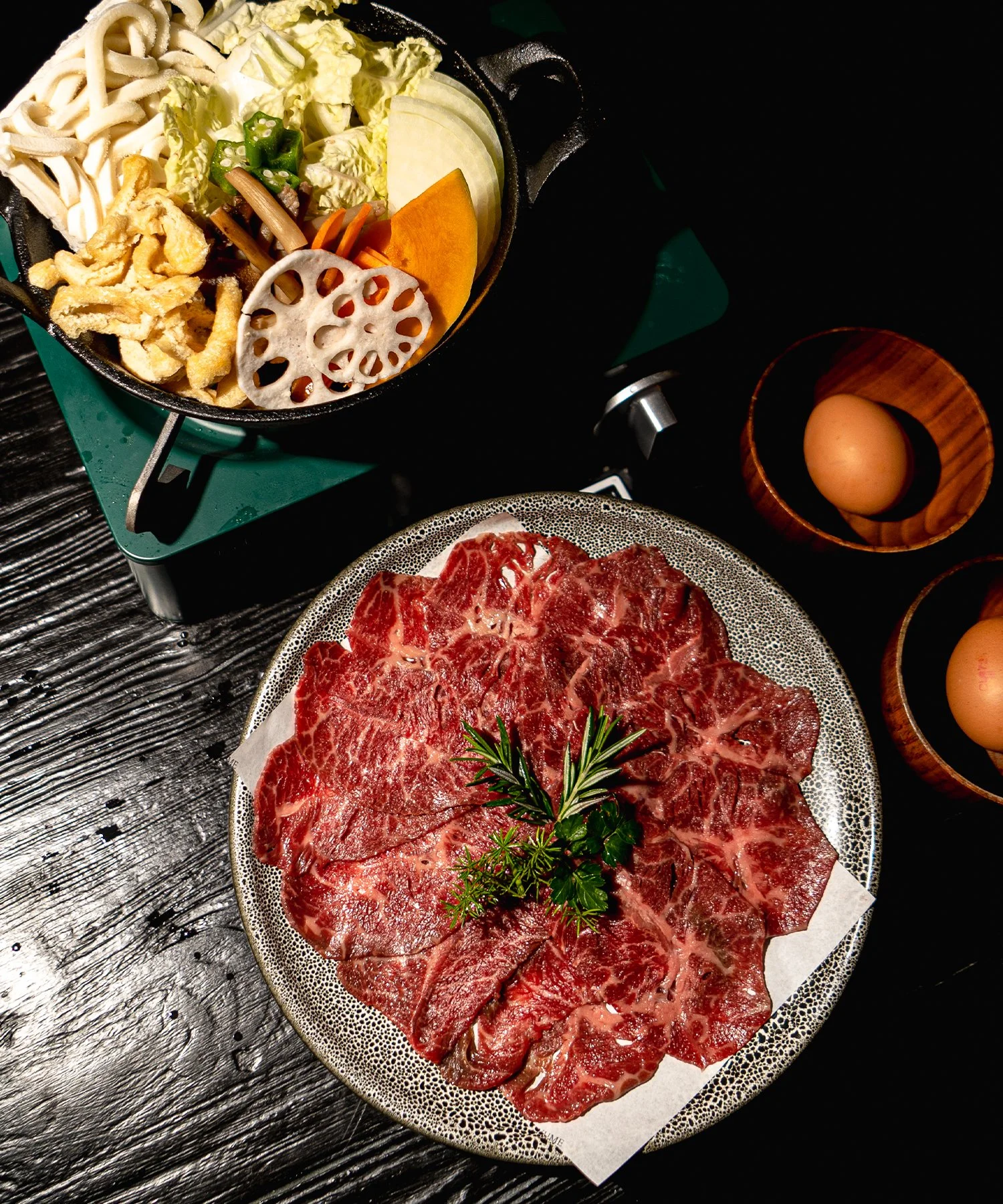 Raw marbled beef slices garnished with herbs, a hot plate with vegetables and noodles, and two eggs in bowls.