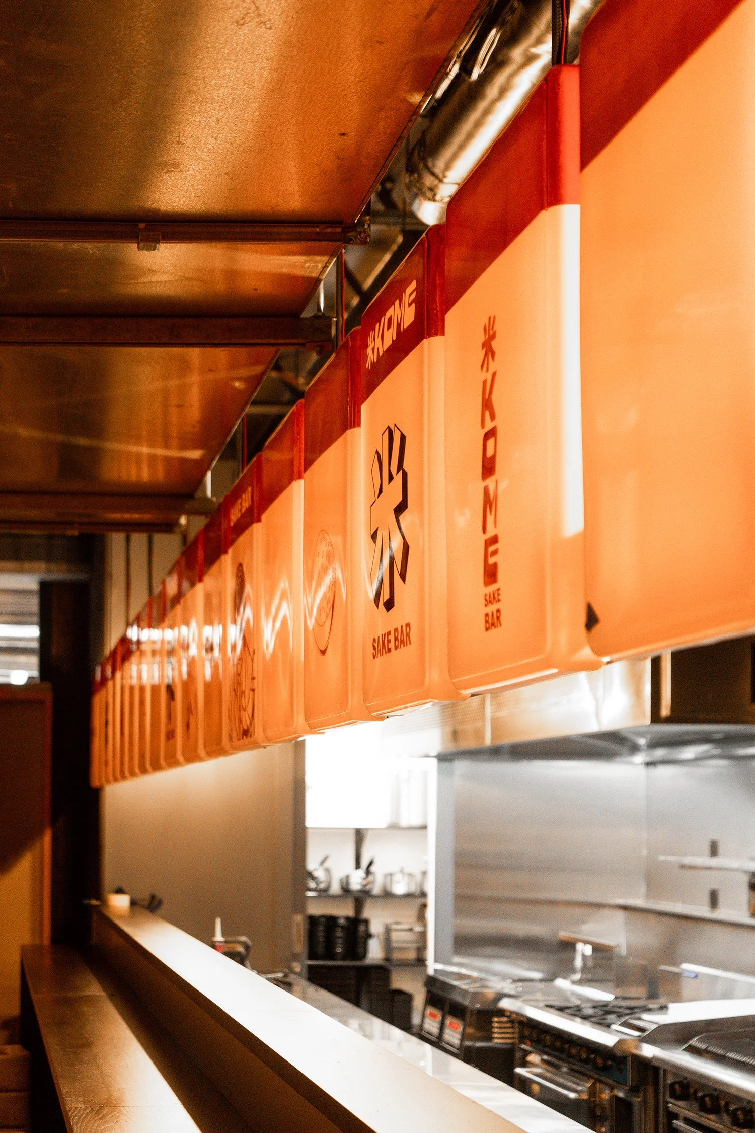 Interior of a bar with orange signs indicating a sake bar, hanging above a counter with kitchen equipment in the background.