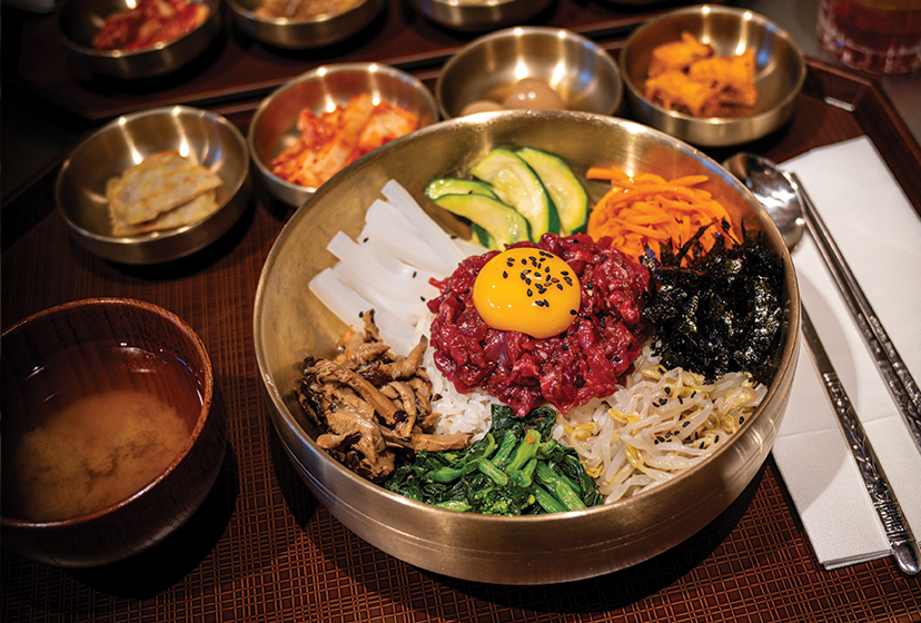 A Korean dish called bibimbap in a metal bowl with raw egg yolk on top, surrounded by various side dishes on a wooden tray.
