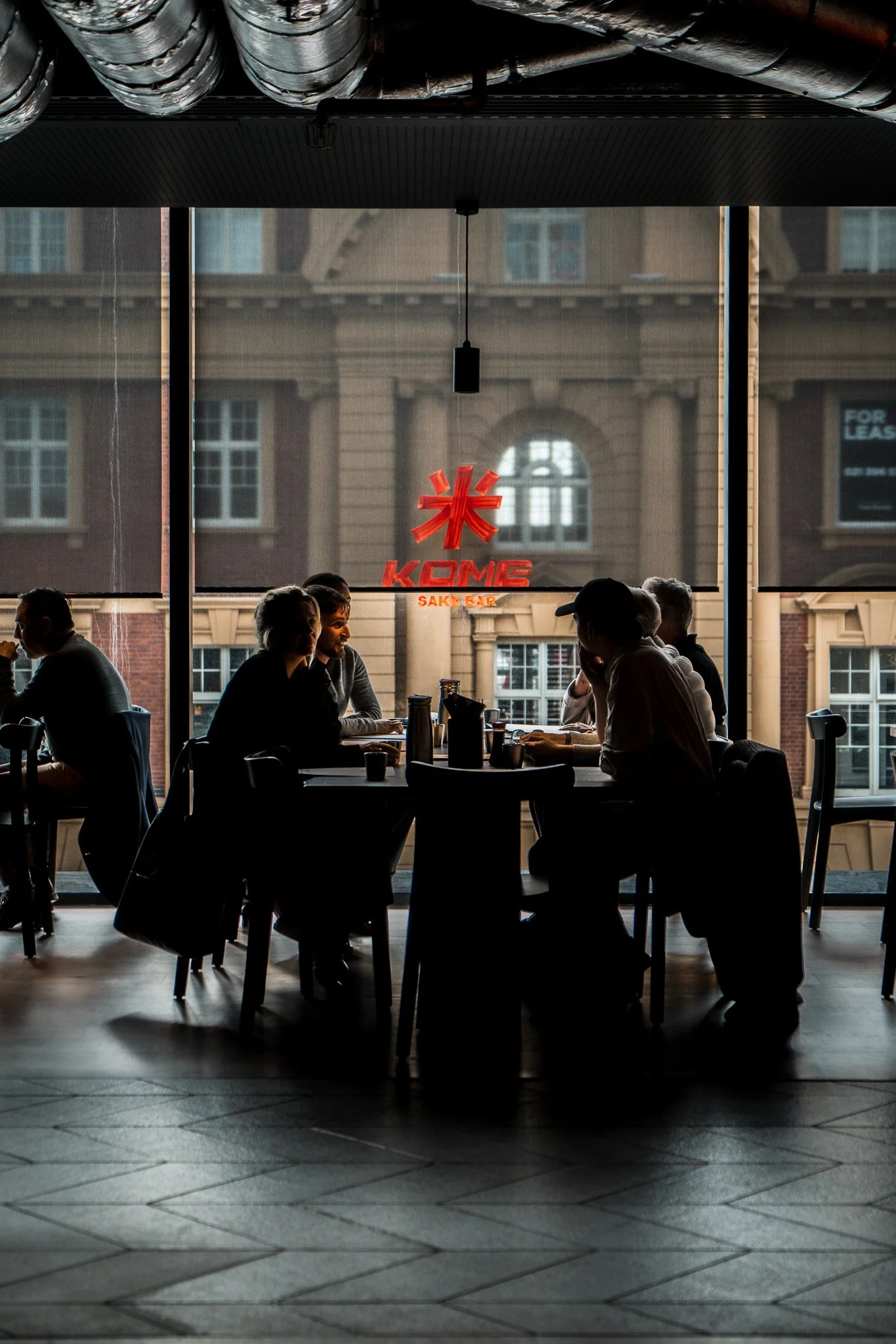 People sitting at a table in a restaurant with large windows showing a city building outside. The restaurant has a sign that says "KOME Sake Bar" in red letters.