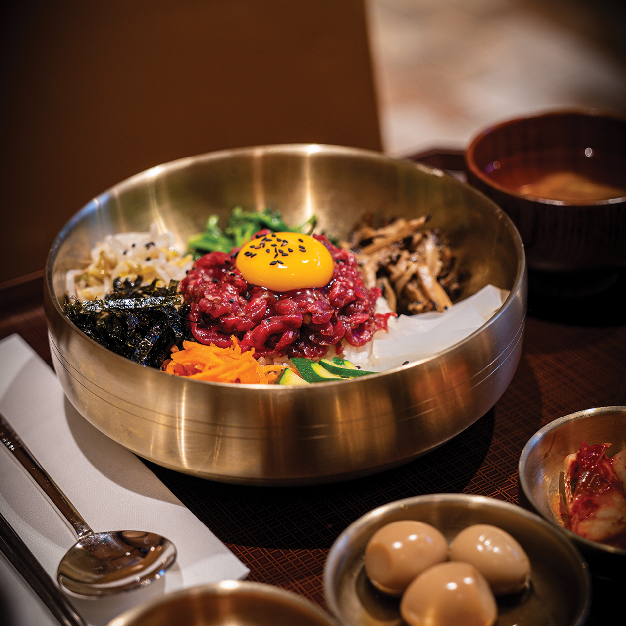 A traditional Korean bibimbap served in a metal bowl with rice, raw beef, egg yolk, and assorted vegetables including carrots, spinach, and seaweed.
