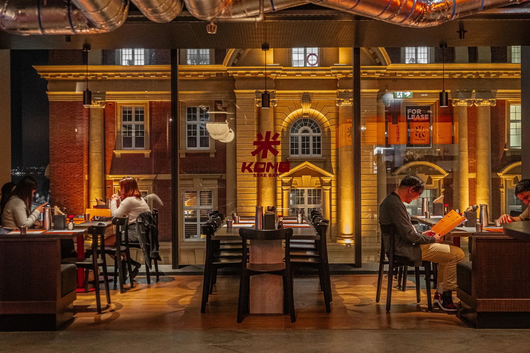 Nighttime scene inside a restaurant with large glass windows showing a historic building's illuminated brick facade outside. The interior has wooden flooring, tables, and chairs, with a few patrons seated, enjoying their meals or reading menus.