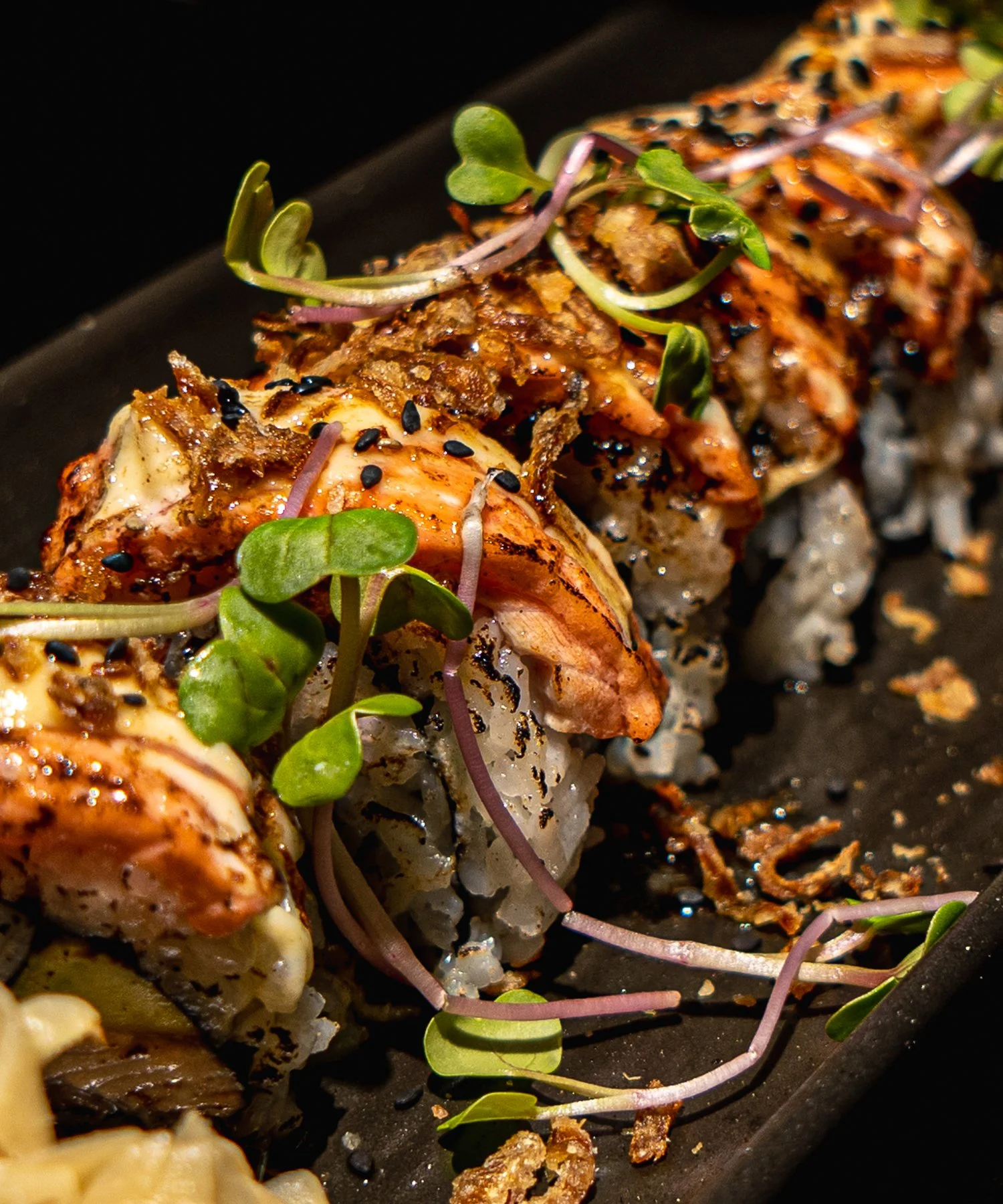 Close-up of a sushi roll topped with cooked seafood, microgreens, black sesame seeds, and drizzled sauce.