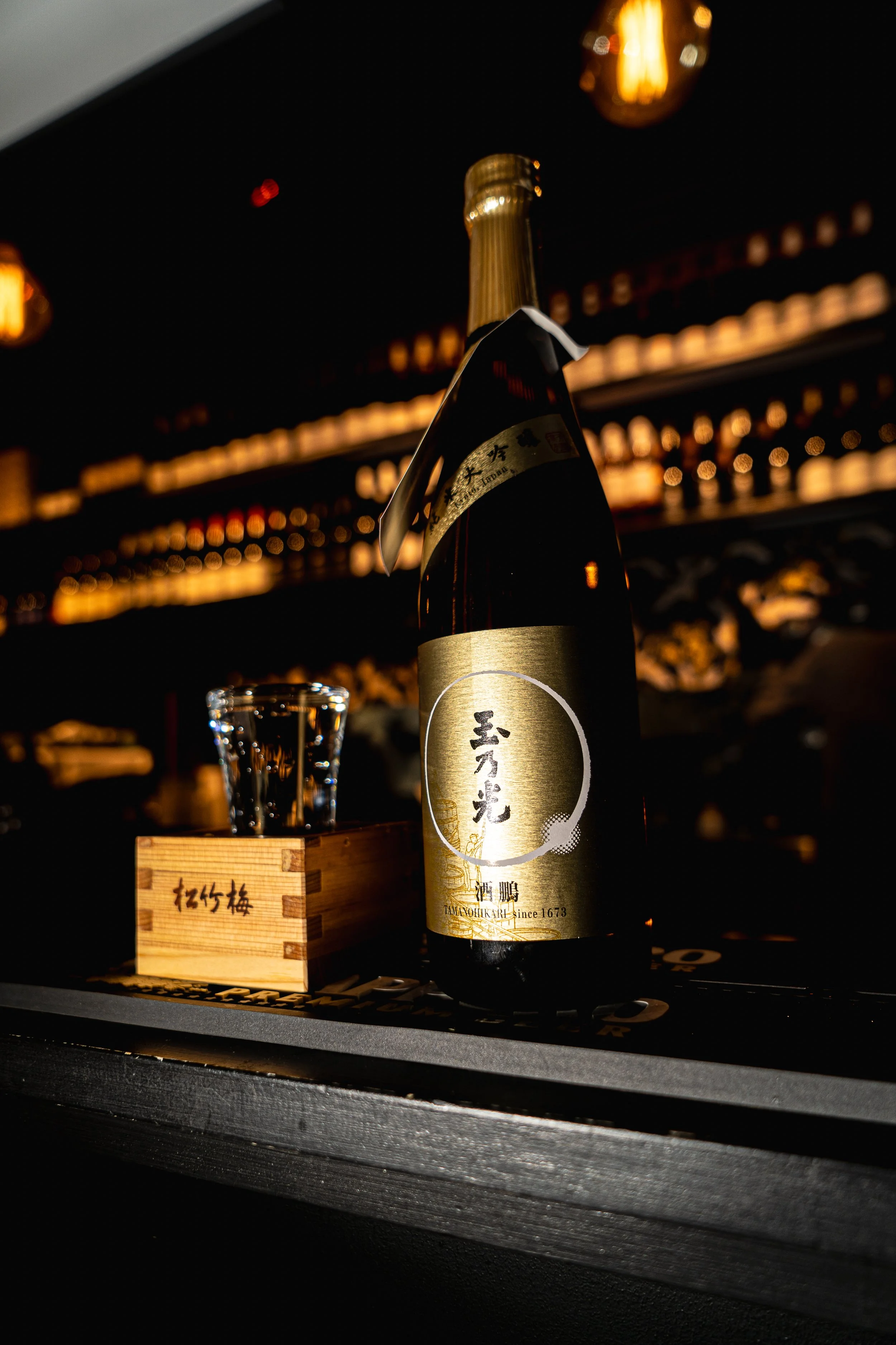 A bottle of Japanese sake on a bar counter, with a small glass and a wooden box beside it, in a dimly lit bar setting.