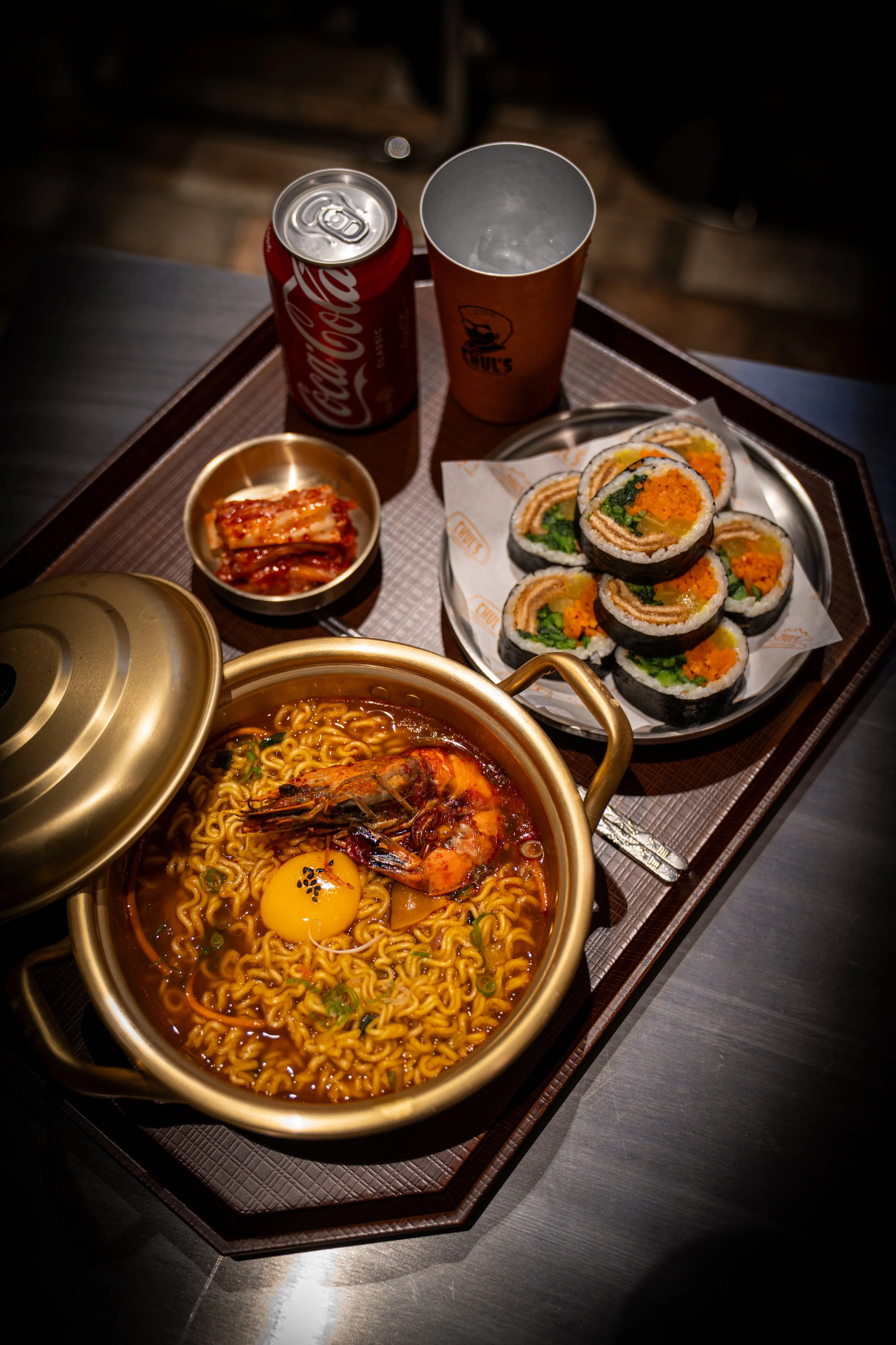 A tray holding a pot of spicy ramen with a shrimp and an egg, a plate of kimbap rolls, a small dish of kimchi, a can of Coca-Cola, a paper cup, and an empty drinking glass.