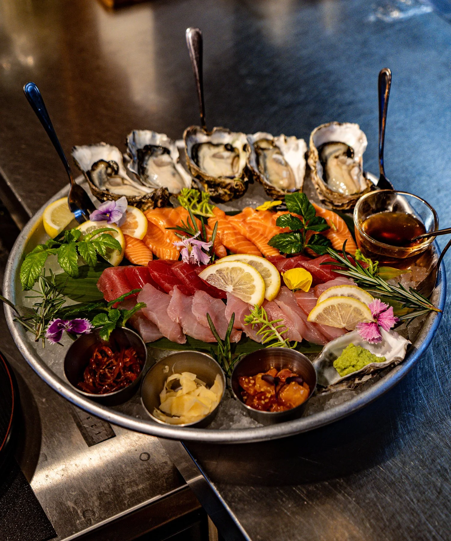 A seafood platter with oysters, salmon, tuna, and other raw fish slices, garnished with lemon slices, herbs, edible flowers, and accompanied by small bowls of dipping sauces and condiments.