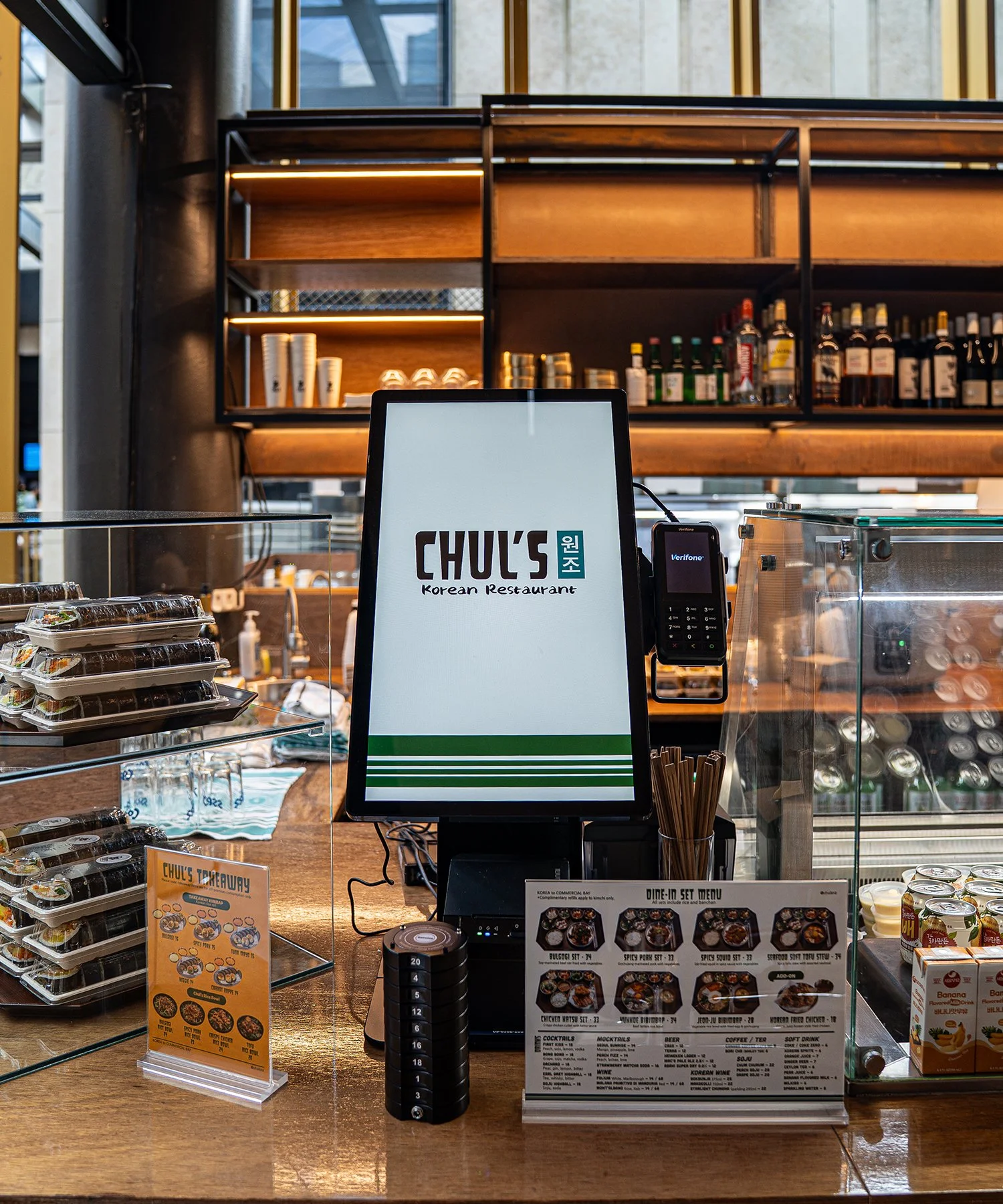 Counter at a Korean restaurant with a digital ordering screen, menu, and food containers, with shelves of bottles and cups in the background