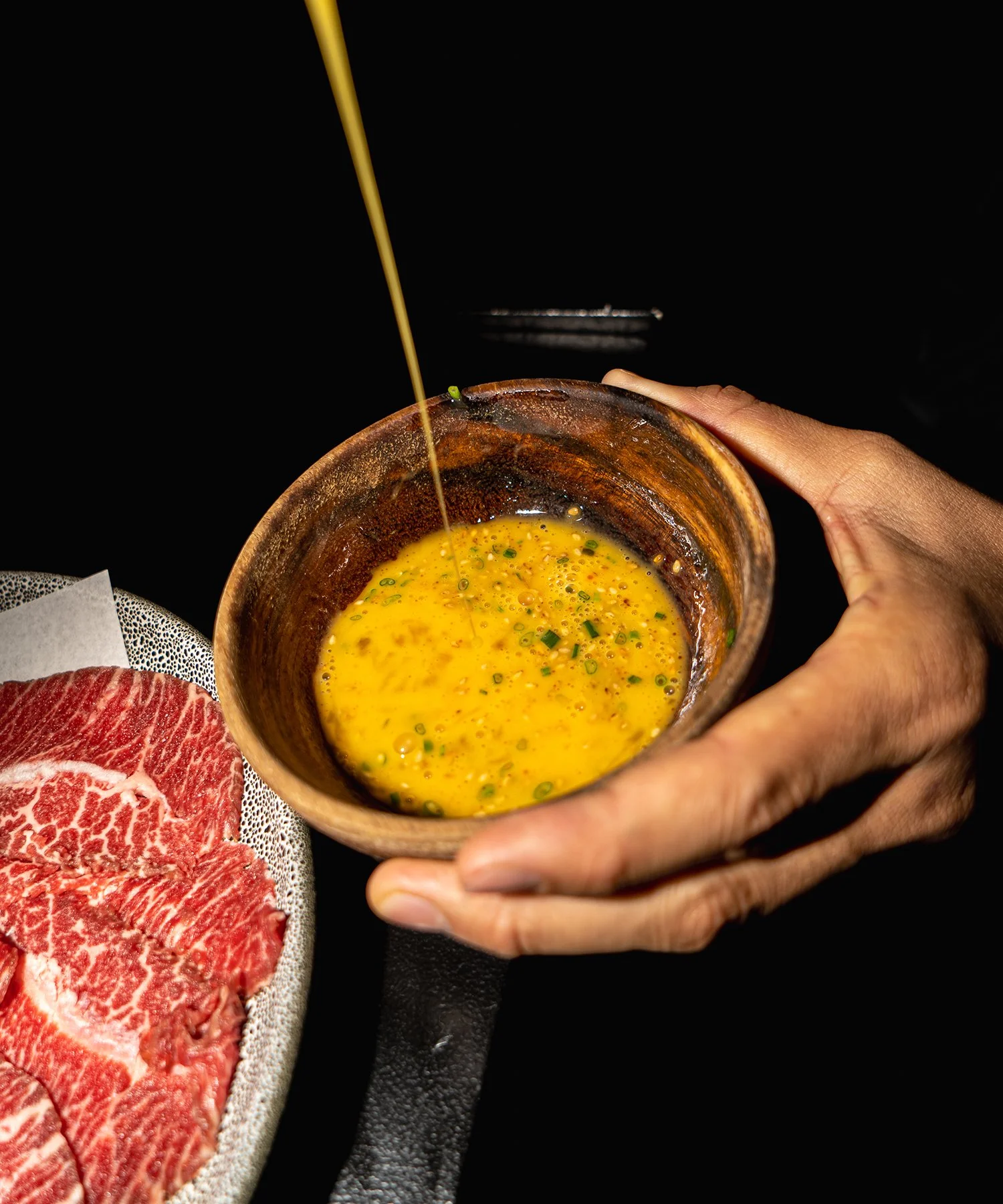 Person holding a small wooden bowl with raw beaten egg, with thin stream of oil pouring into it, next to a plate of thinly sliced raw beef.