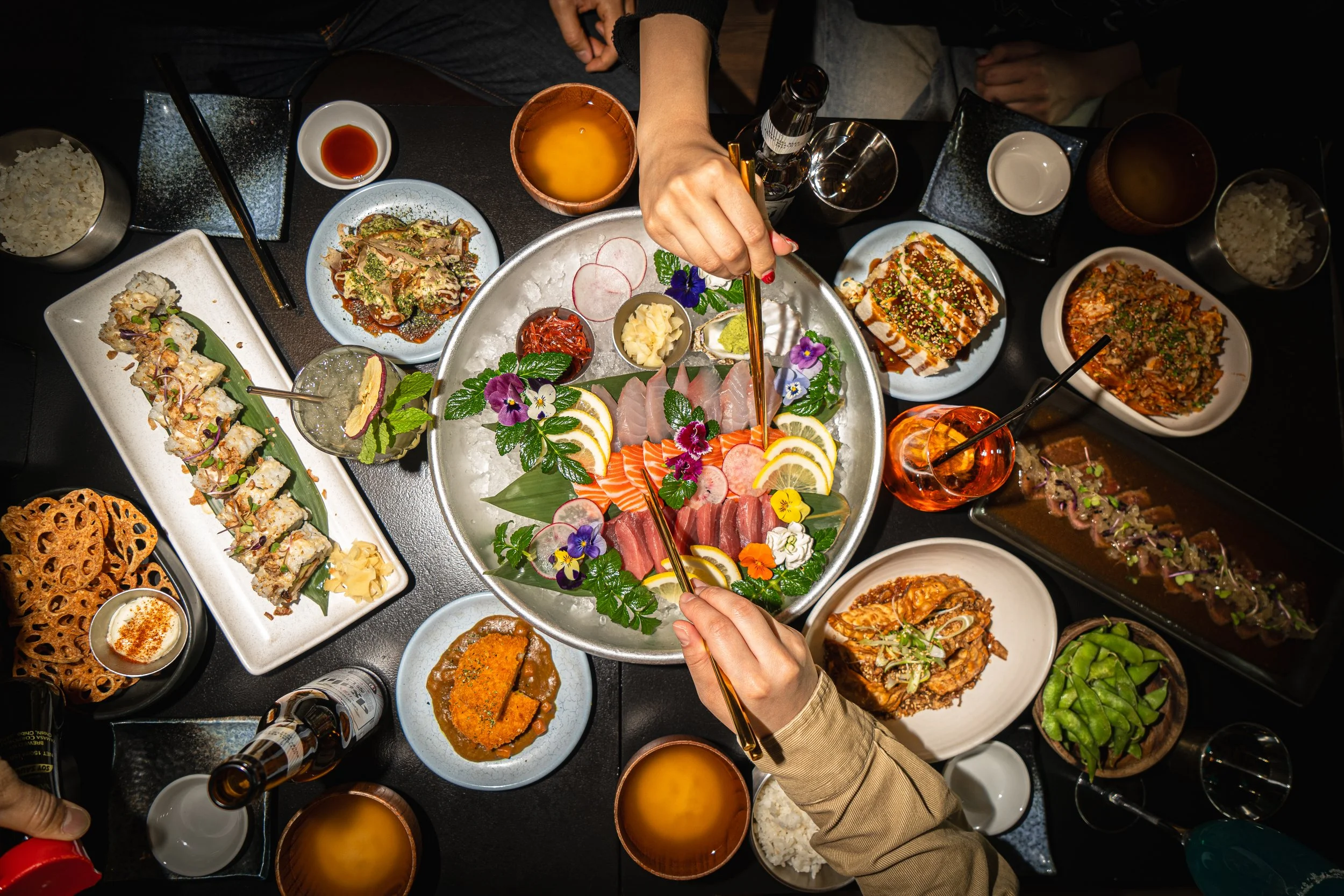 A top-down view of a Japanese sushi dinner with various dishes, sashimi, alcohol, and sides on a black table.