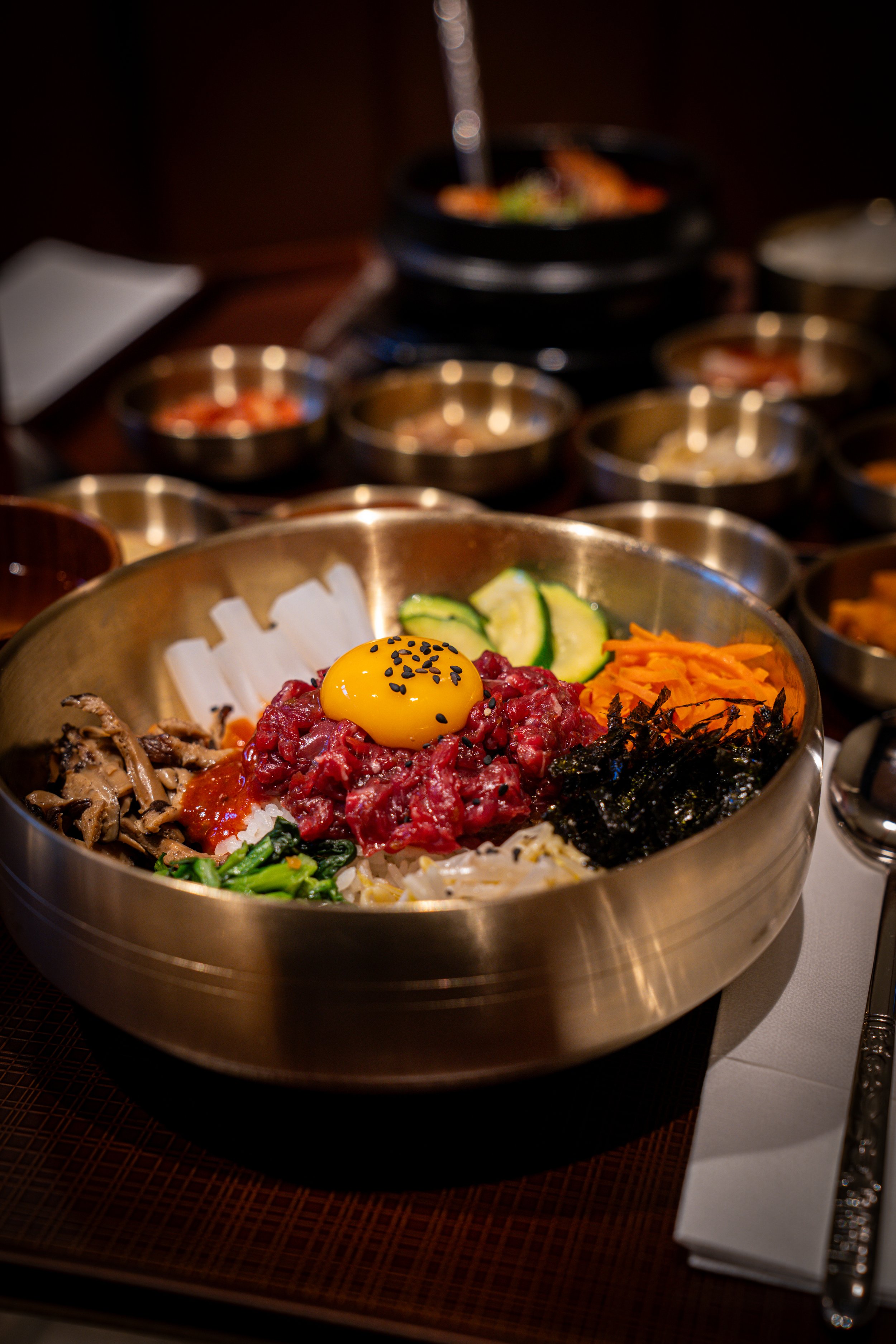 Bowl of Korean bibimbap with raw egg yolk, vegetables, and sliced beef, on a table with small side dishes.