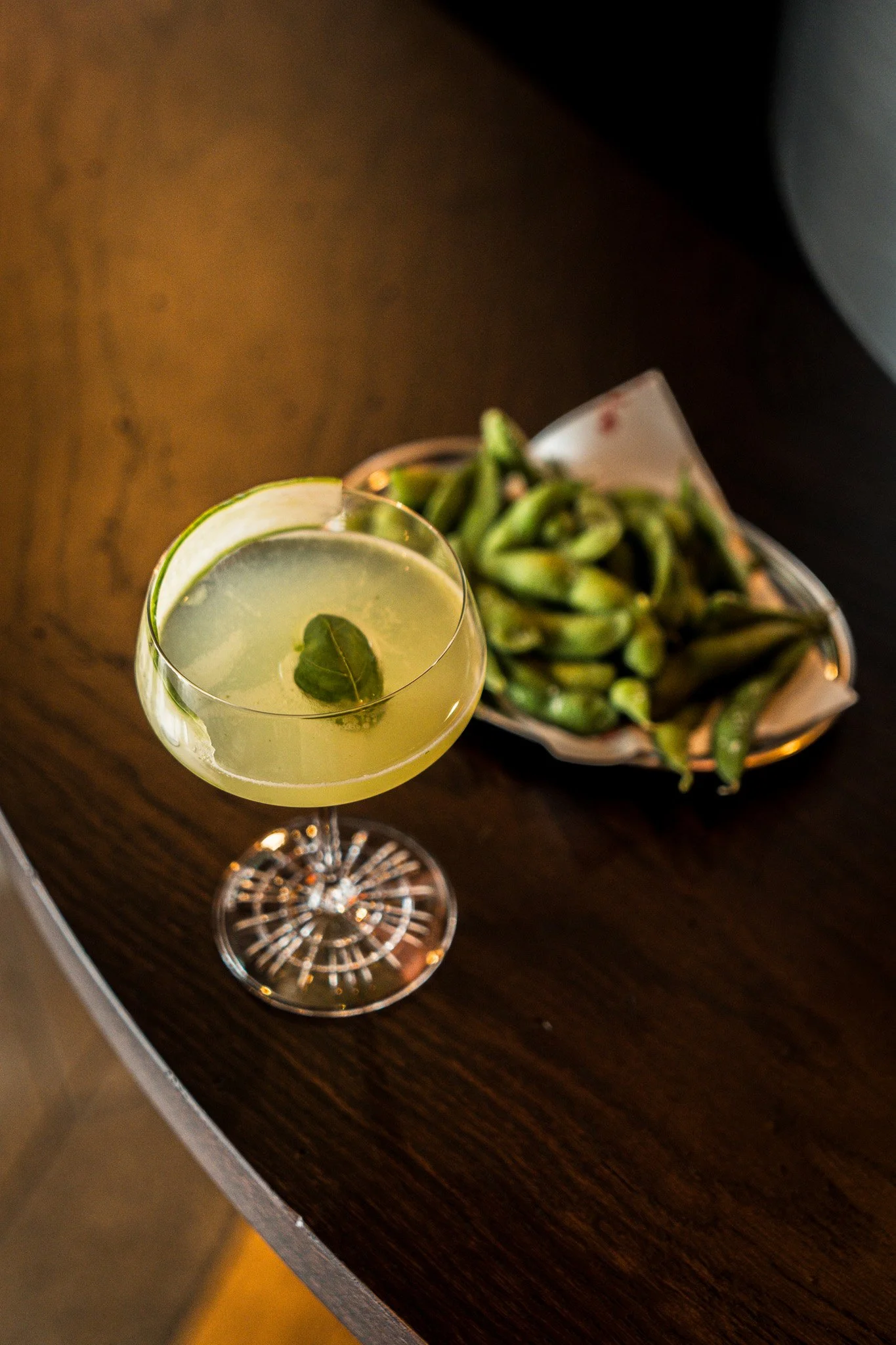 A cocktail in a fancy glass garnished with a lime wedge and a leaf, placed on a wooden table next to a bowl of edamame beans.