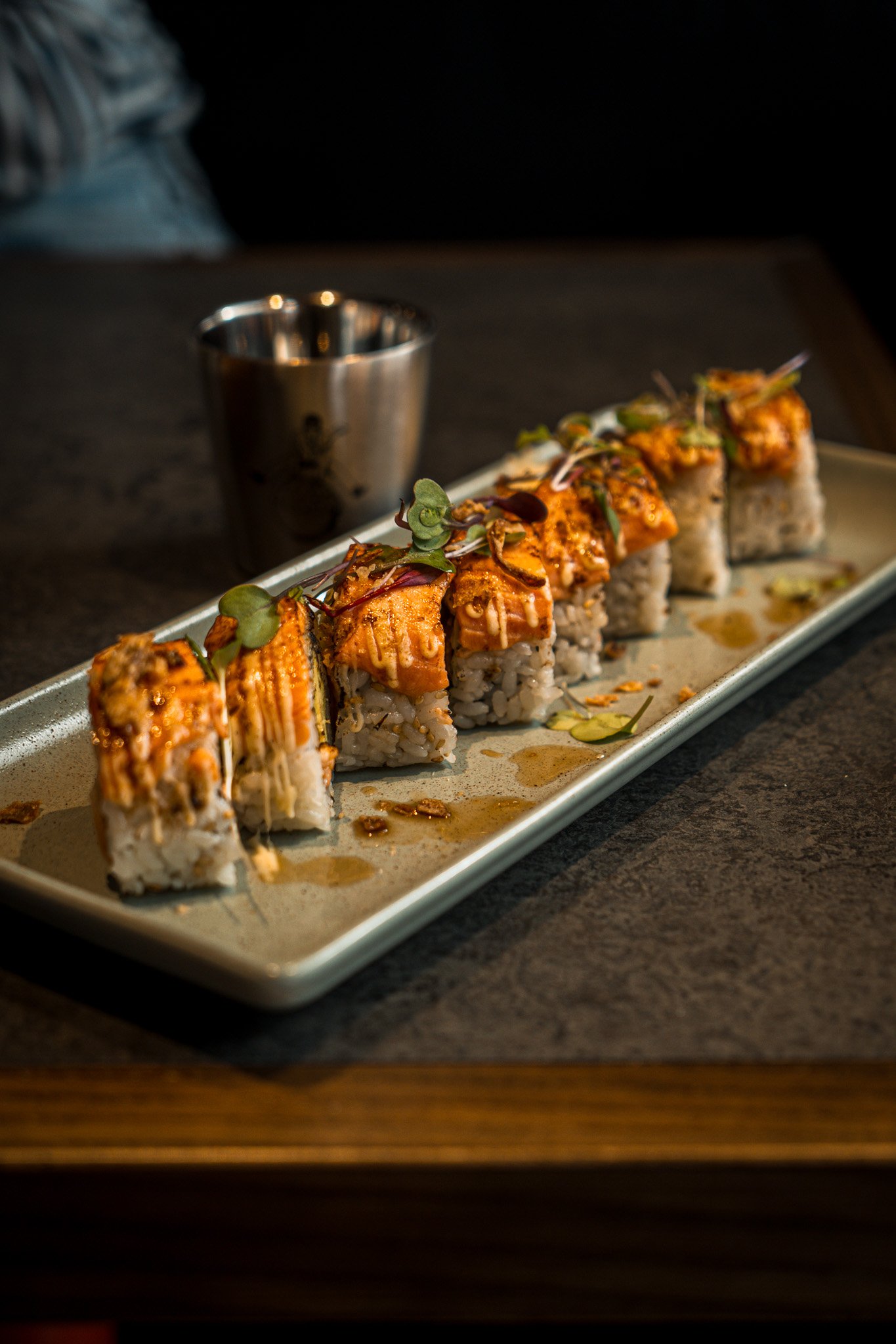Sushi rolls topped with eel, garnished with microgreens on a rectangular plate, with a small metal cup in the background.