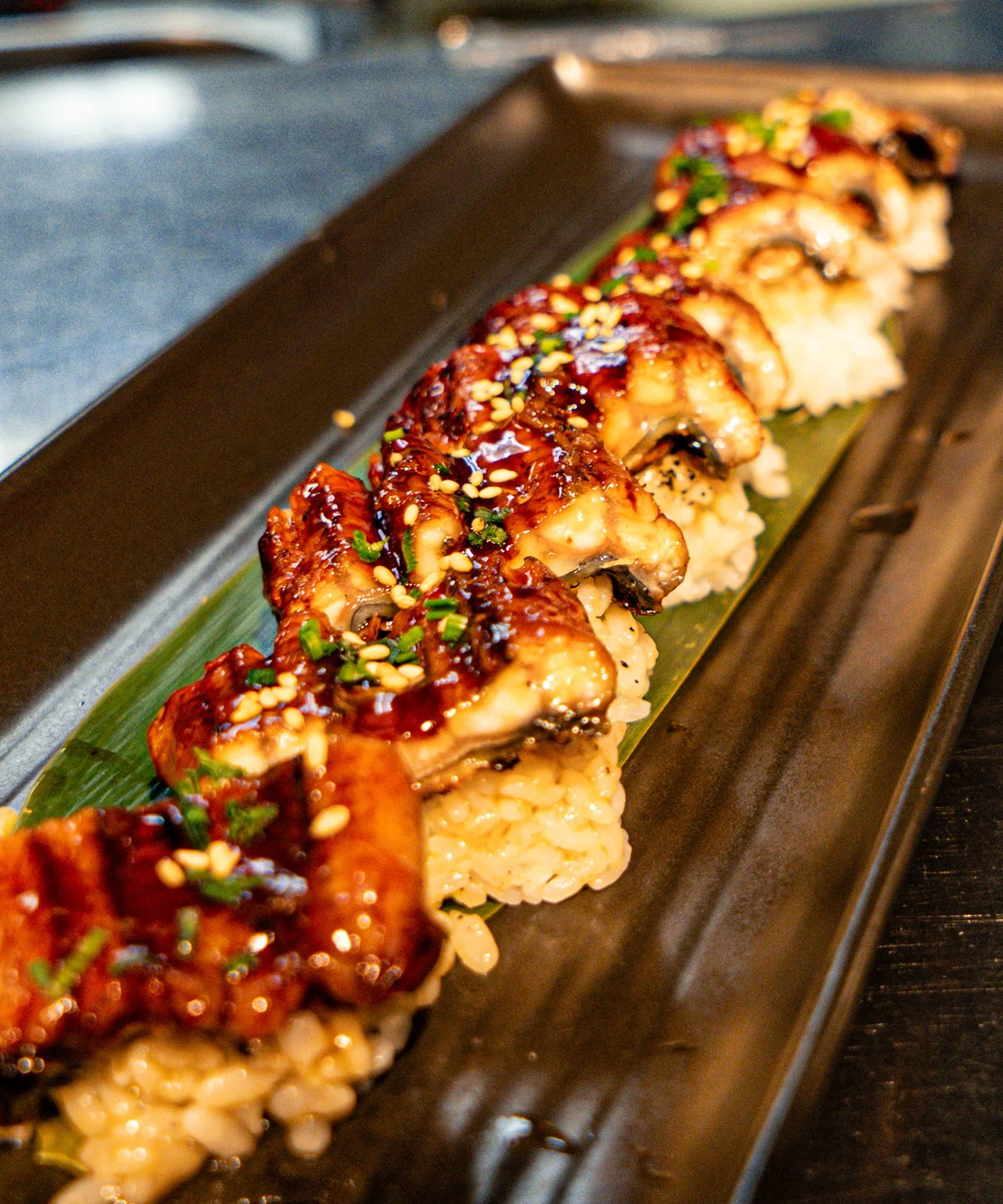 Sushi rolls topped with eel sauce, sesame seeds, and chopped green onions on a black rectangular plate with a green leaf.