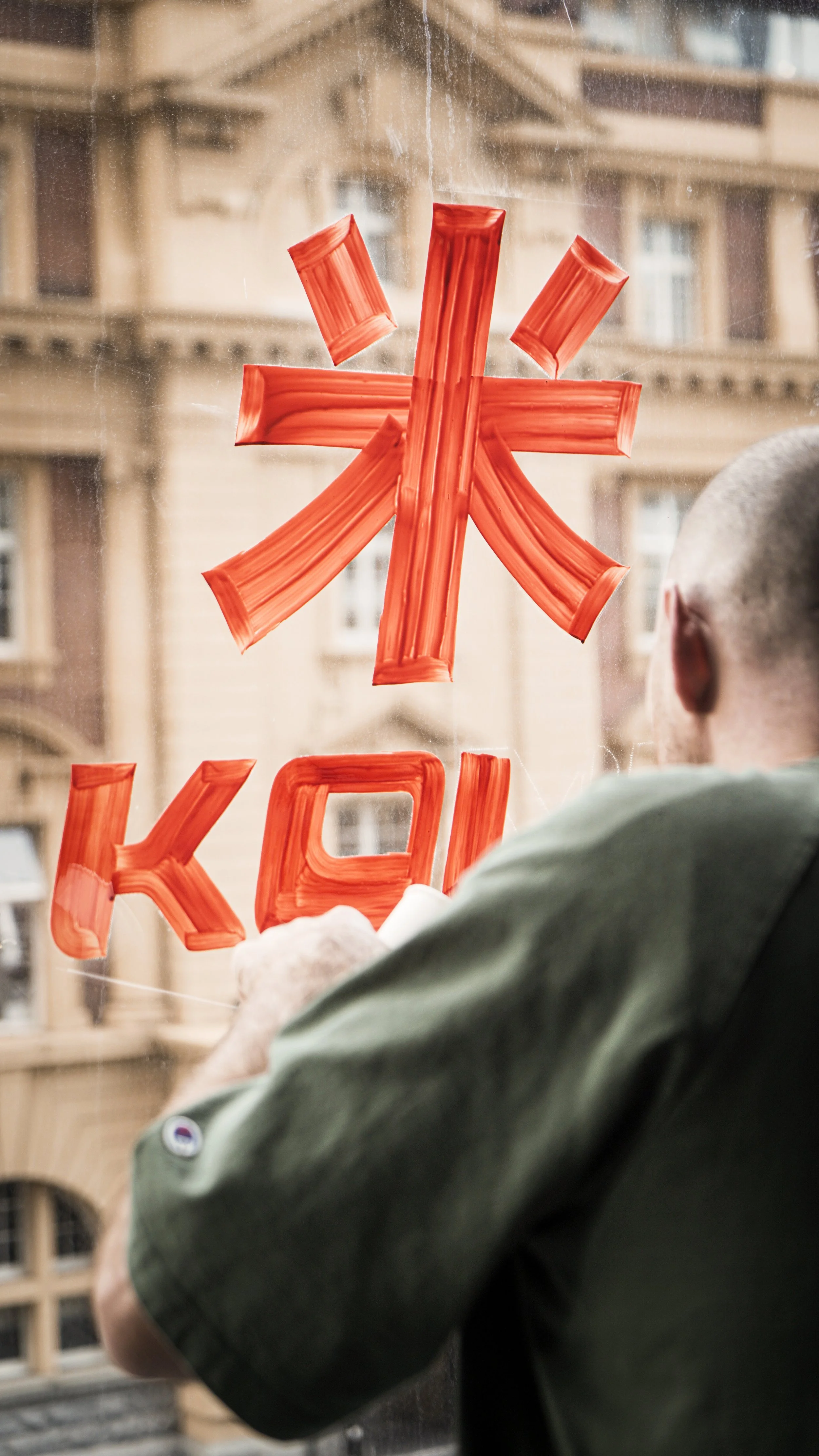 A person is painting red symbols and letters on a glass window, with an building visible in the background. The symbols include a stylized asterisk and the letters 'K' and 'A'.