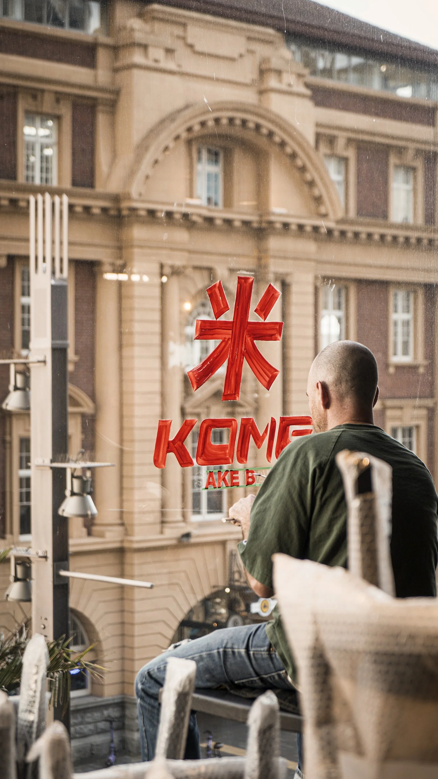 A man sitting on a balcony, looking out a glass window with a red sign that reads 'KOME' and a symbol above it, with a historic building visible outside.