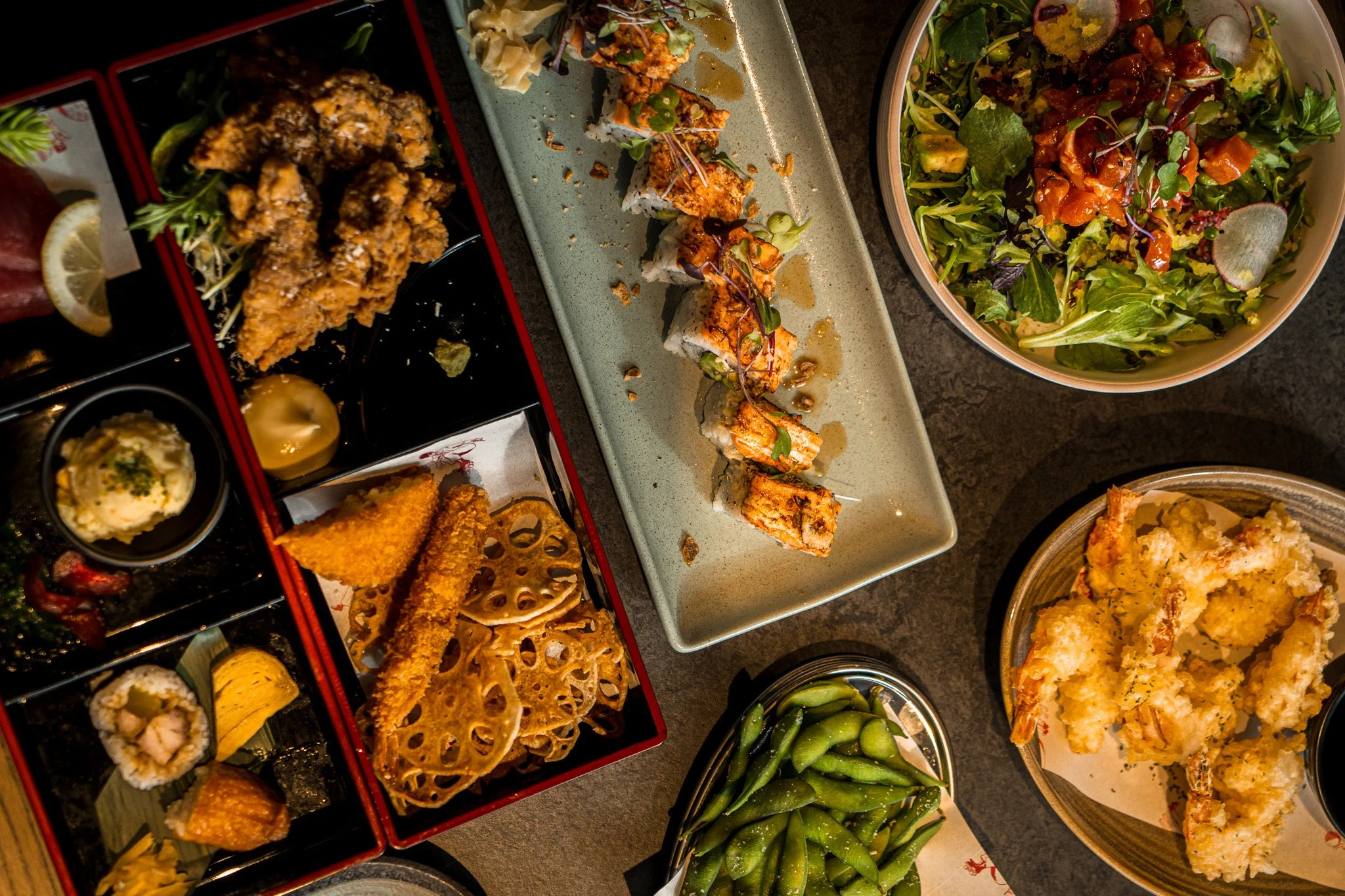 A variety of Japanese cuisine dishes, including sushi rolls, tempura, edamame, salad, and fried chicken, arranged on a table.