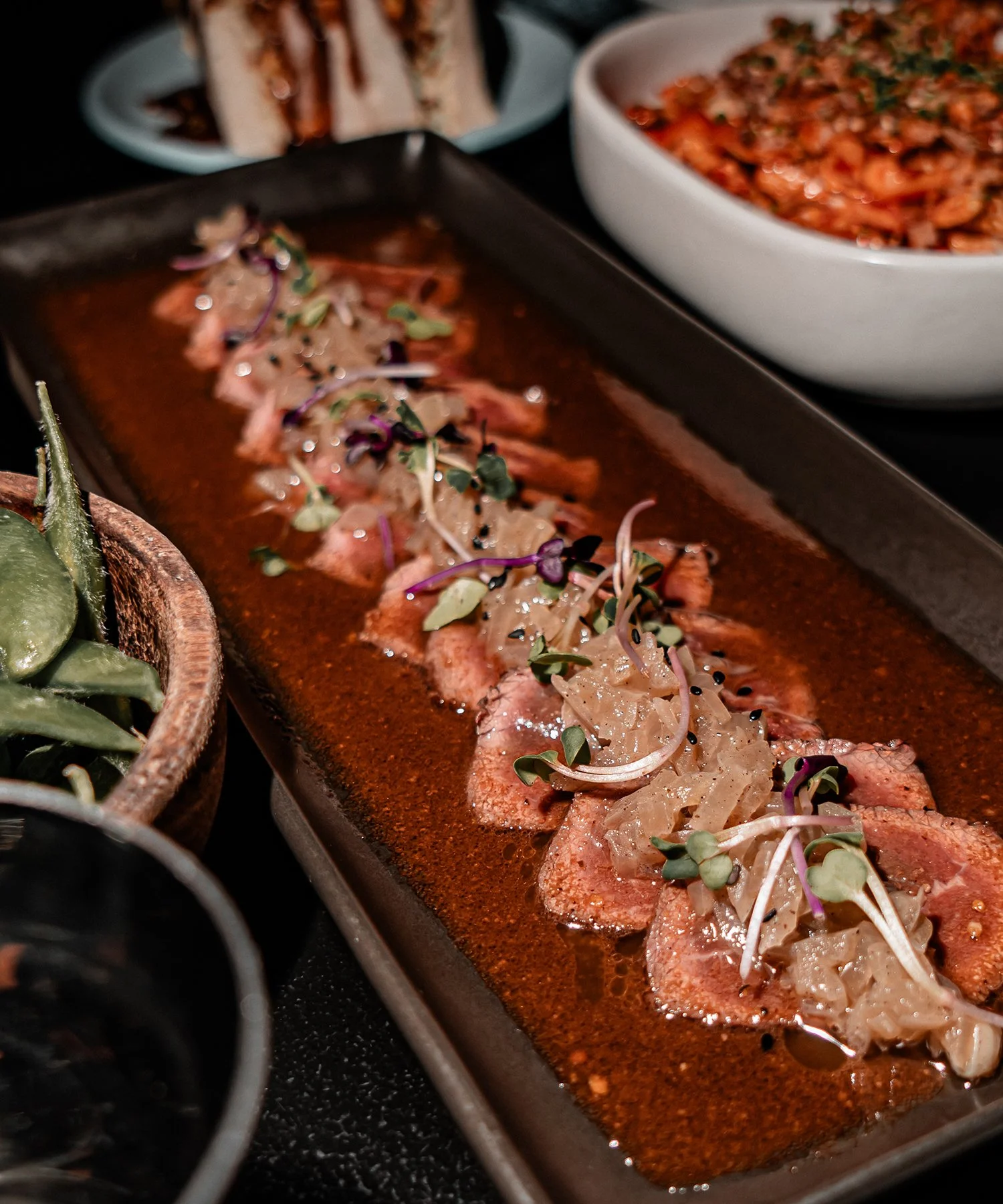 A row of sliced, cooked meat topped with caramelized onions, microgreens, and black sesame seeds, served on a rectangular black plate with various side dishes in bowls around it.