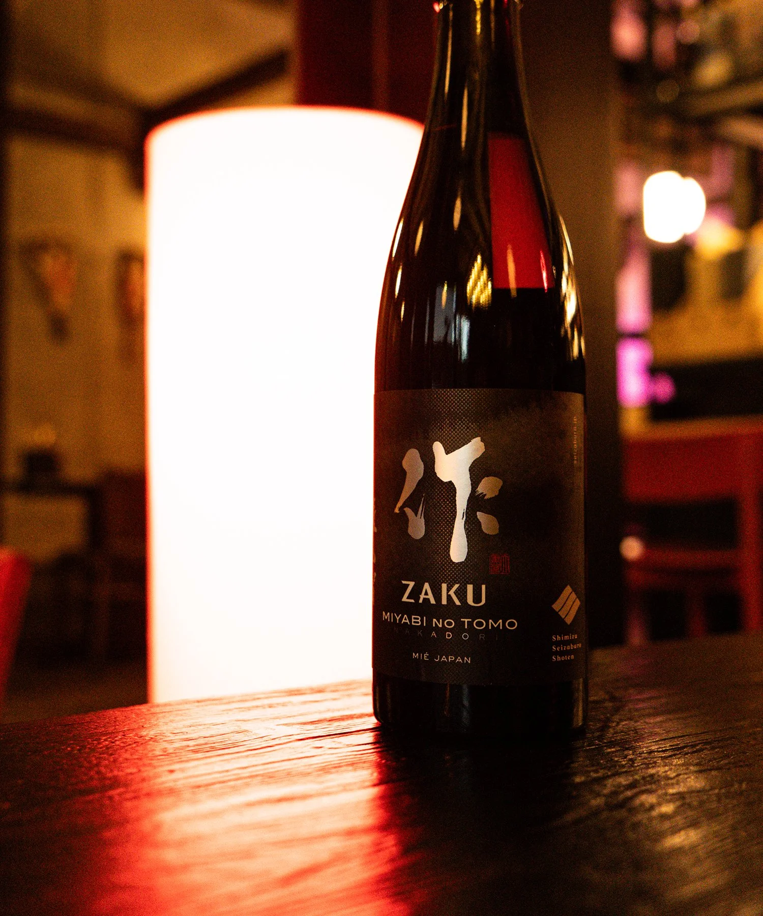 A bottle of Japanese sake on a wooden table, illuminated by a nearby light, in a restaurant or bar setting.
