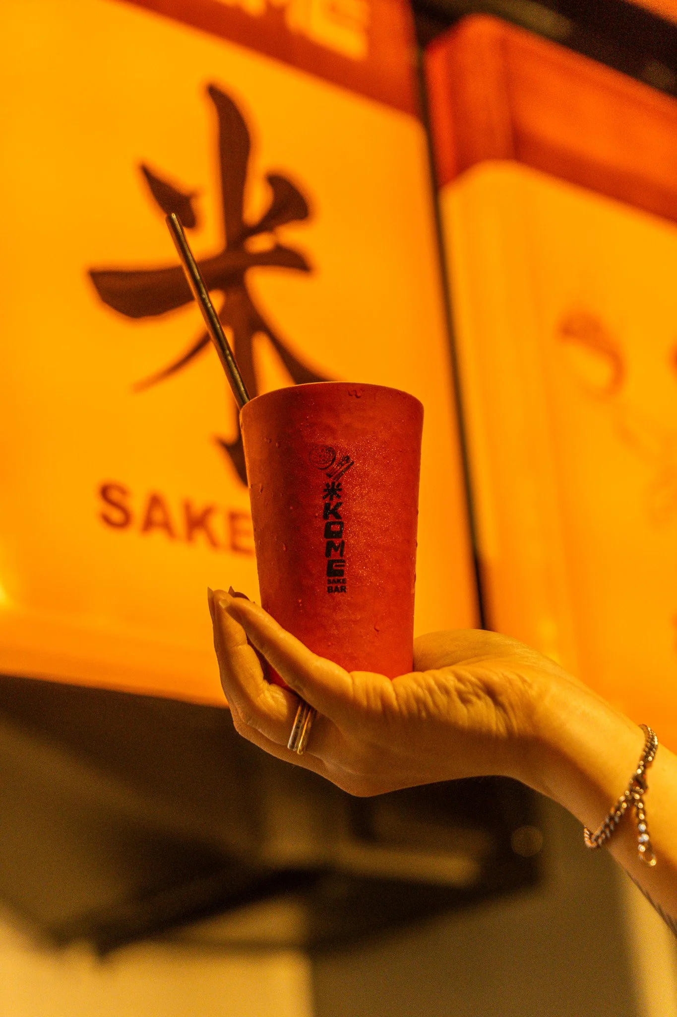 A hand holding a red cup with a metallic straw at a sake bar, with a large illuminated Japanese character on the yellow background behind.