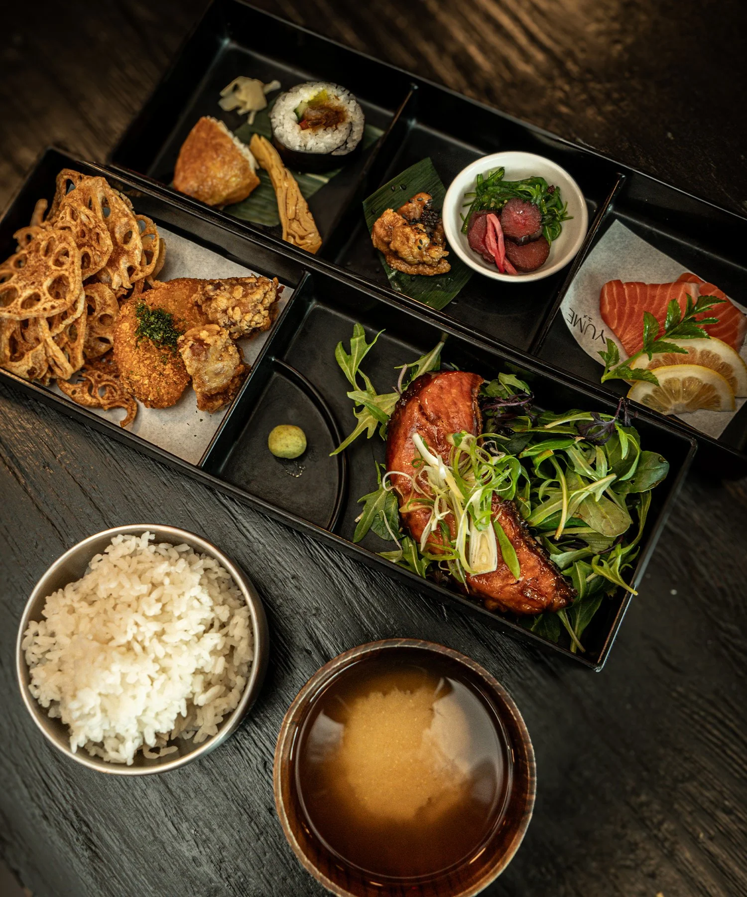 A traditional Japanese bento box meal with rice, miso soup, and assorted small dishes including sushi, tempura, grilled fish, salad, pickles, and lemon slices.