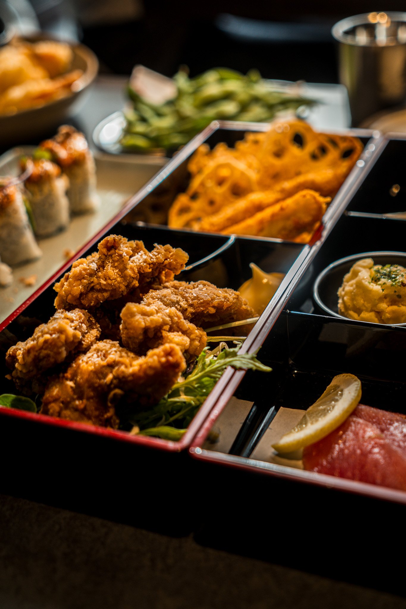 A bento box with fried chicken, spring rolls, and various side dishes including mashed potatoes and vegetables, garnished with lemon slices.