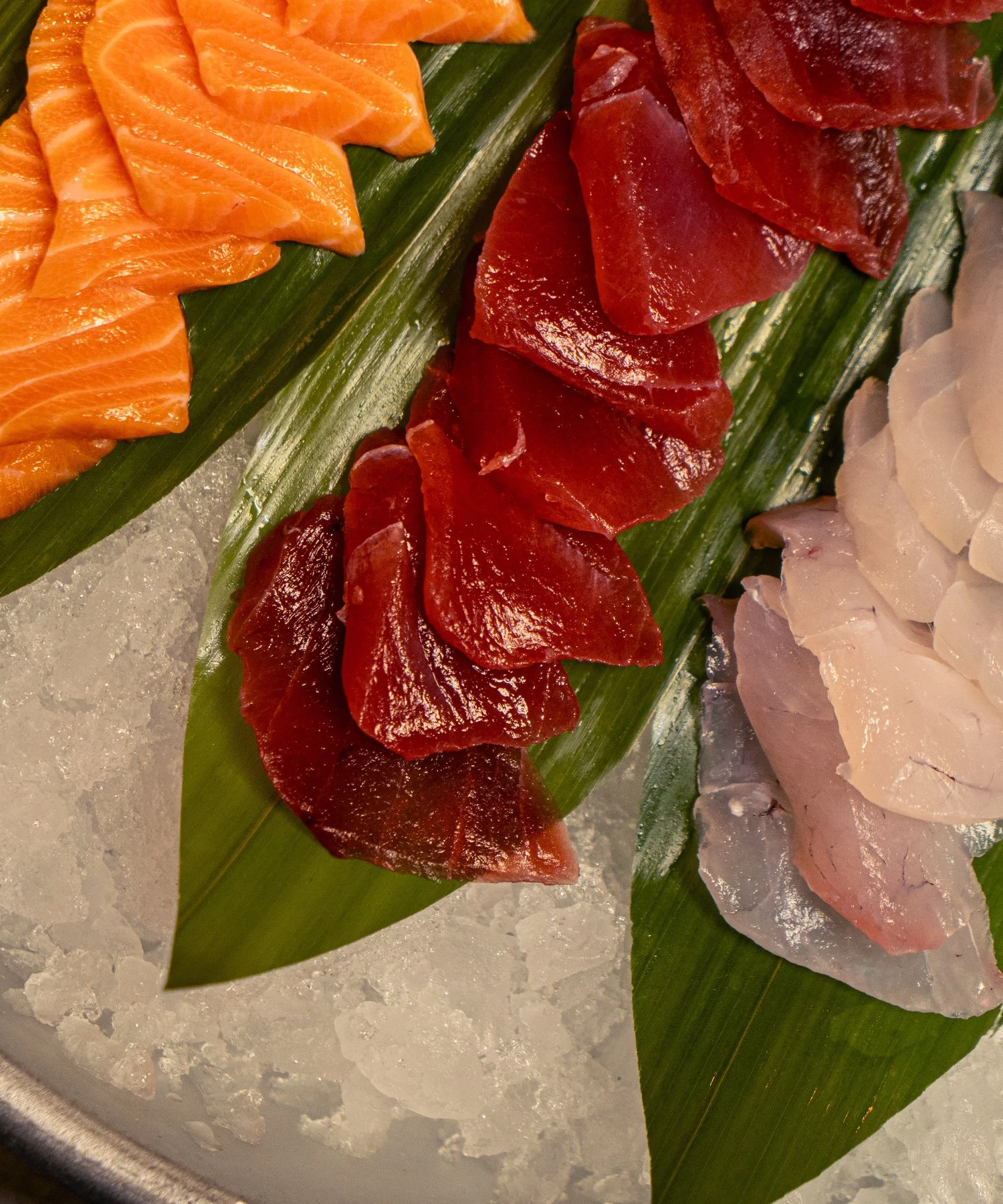 Sashimi slices of salmon, tuna, and whitefish arranged on green leaves over ice.