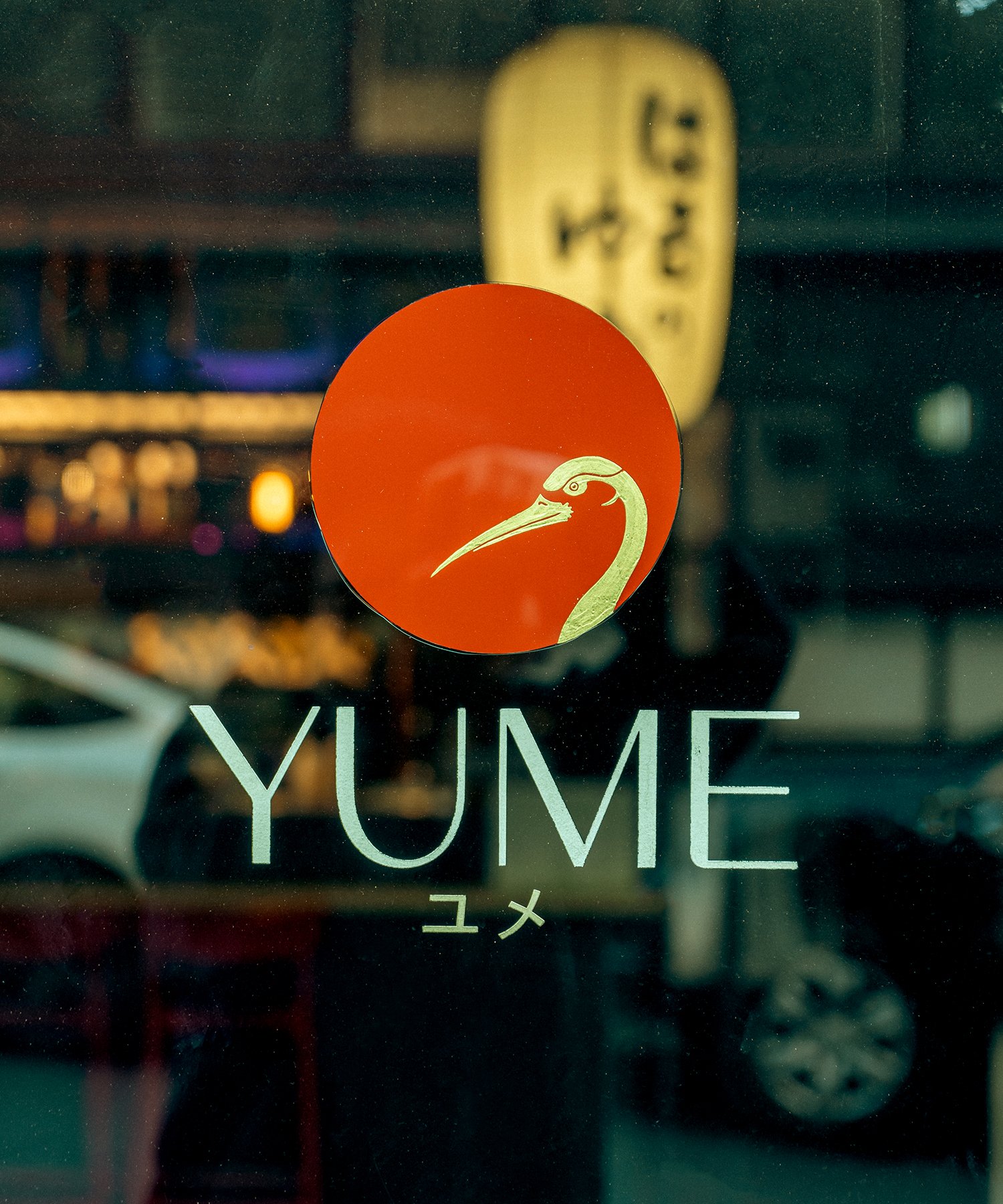 A glass window with the logo of a restaurant called YUME, featuring a heron or crane bird illustration inside a red circle, and Japanese characters below the name.