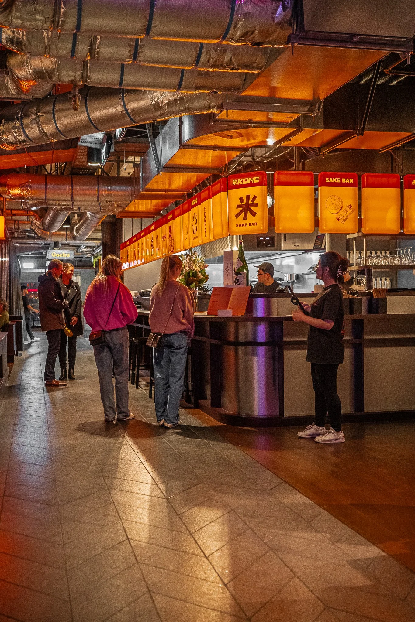 People line up at a sushi bar inside a restaurant, with a kitchen visible in the background and illuminated signs displaying 'Sake Bar' and Japanese symbols.