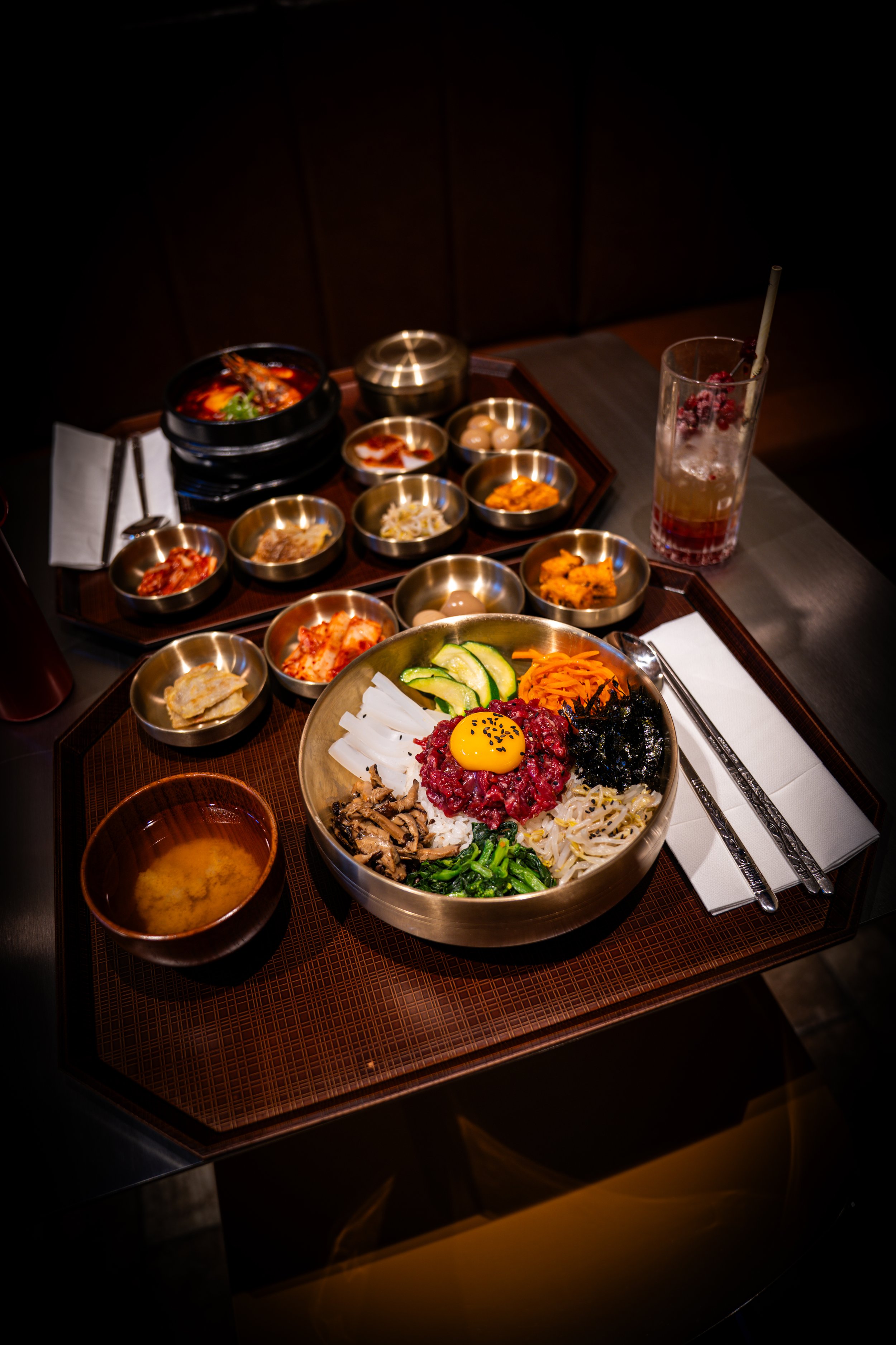 A traditional Korean bibimbap dish with a raw egg yolk on top, surrounded by various side dishes and a bowl of soup.