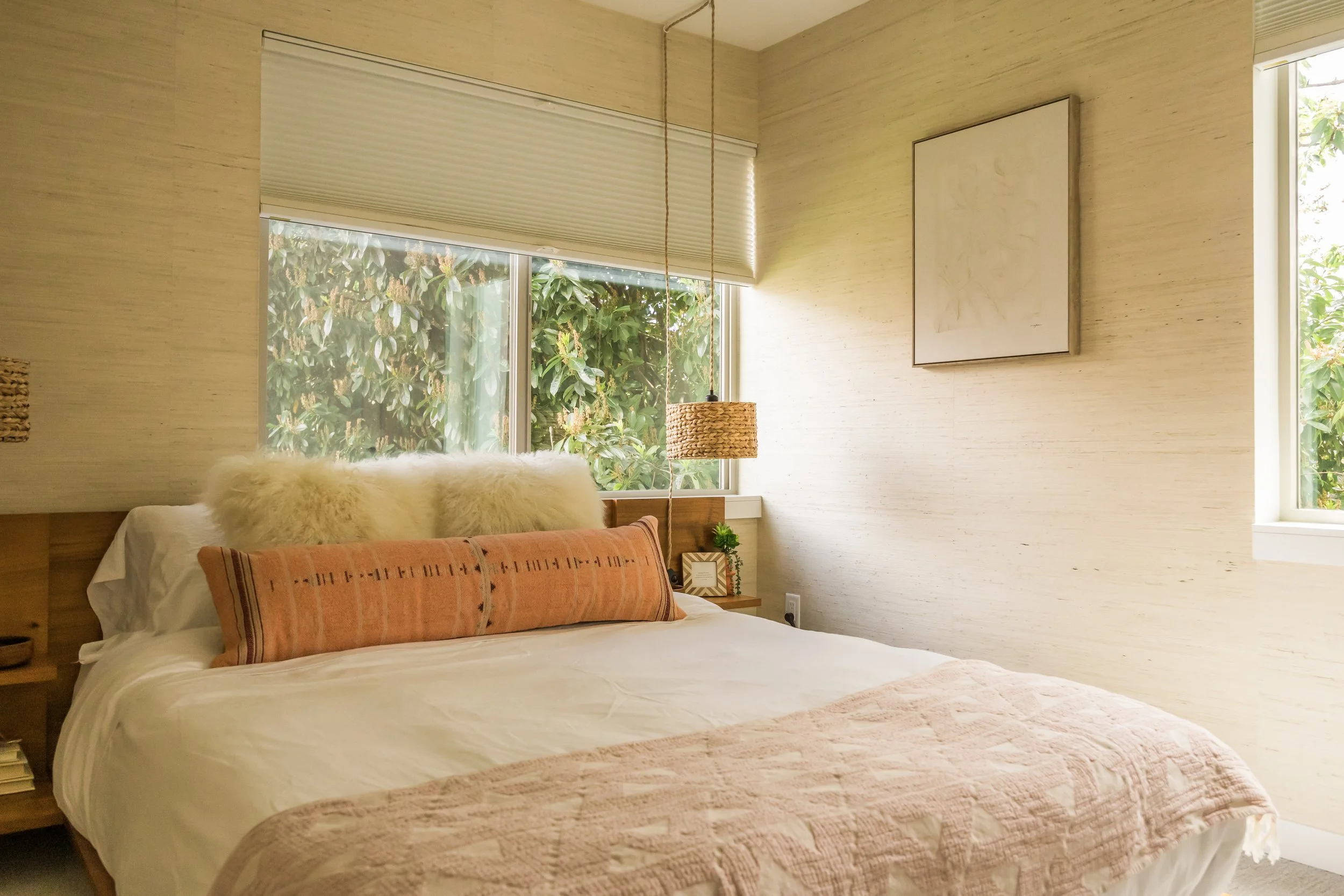 Bright bedroom with beige walls, large windows with greenery outside, a bed with white bedding, a long orange pillow, and fluffy white pillows at the head, with a woven pendant light hanging above and a small bedside table with a plant.