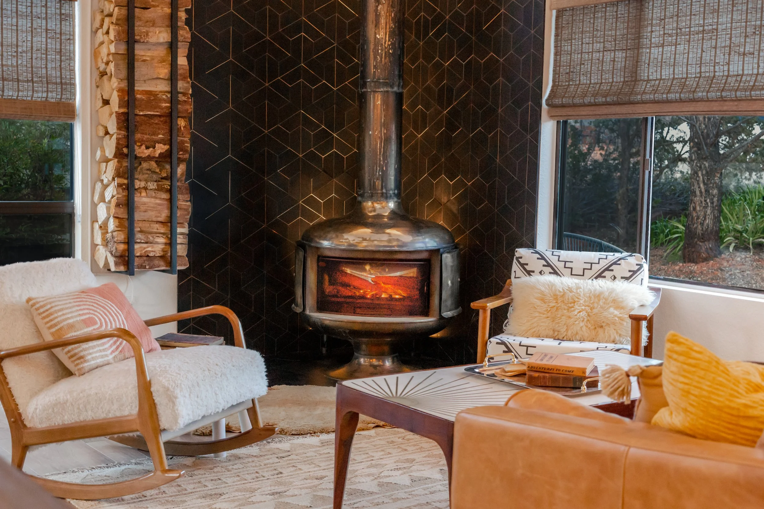 Living room with a black fireplace, two windows with blinds, two chairs with pillows, a coffee table with books and tray, and a tan sofa with cushions.