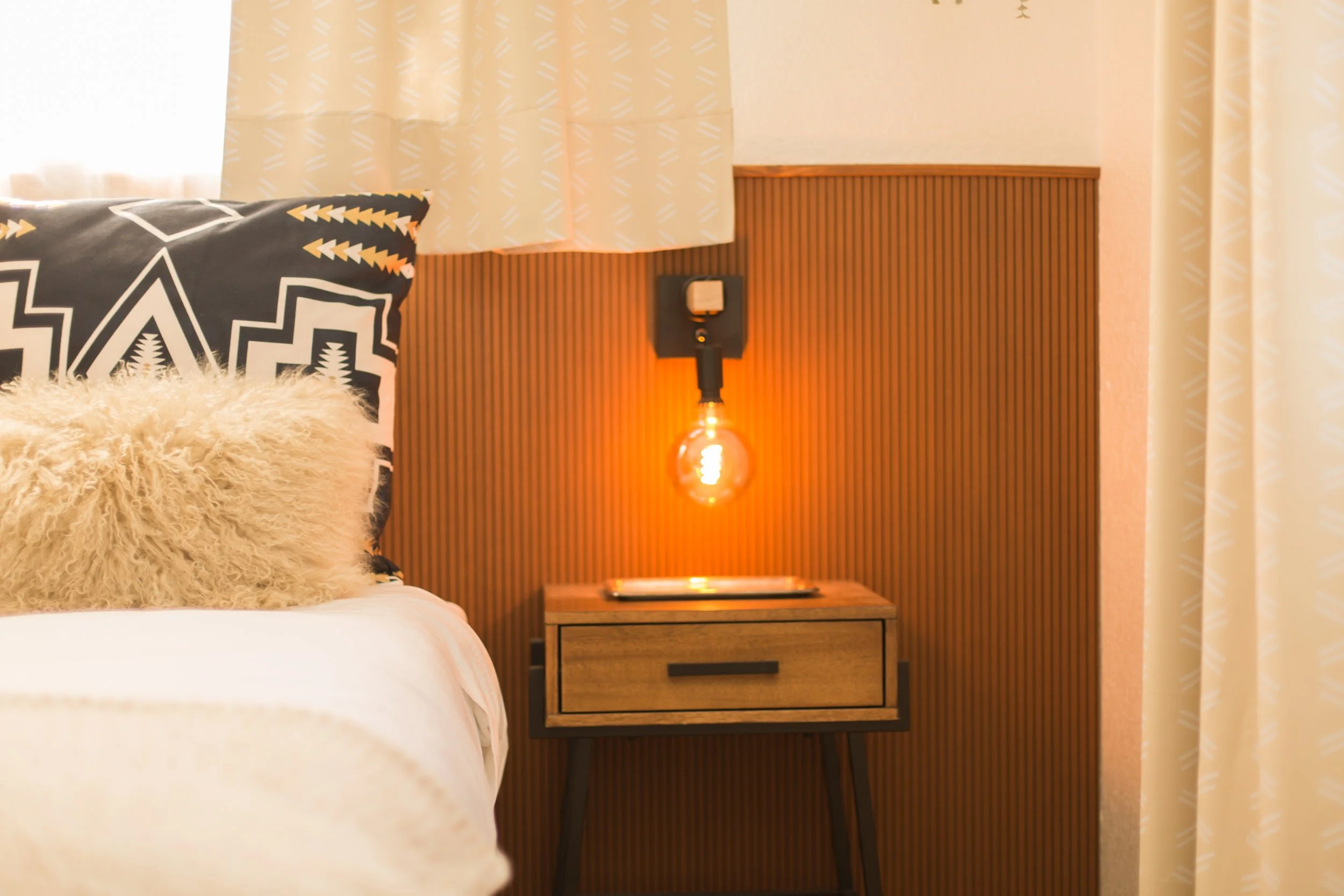 A bedroom corner with a wooden nightstand, a warm orange-glowing hanging bulb, a furry beige pillow, and patterned black and white pillows on a bed, with cream curtains covering a window.
