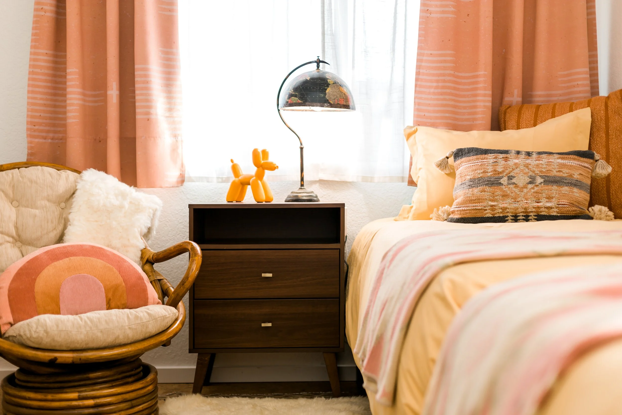 A bedroom with pink curtains, a bed with yellow and pink bedding, a decorative pillow, a wooden side table with a modern desk lamp, a yellow balloon dog sculpture, and a rattan chair with a rainbow pillow.