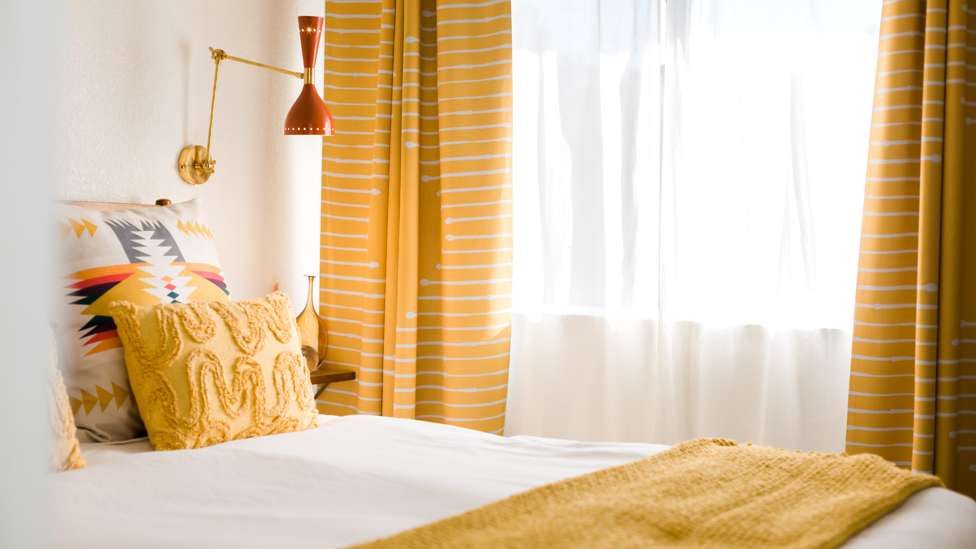 A cozy bedroom with yellow and white striped curtains, a white bed with patterned and textured yellow pillows, and a red wall-mounted lamp.