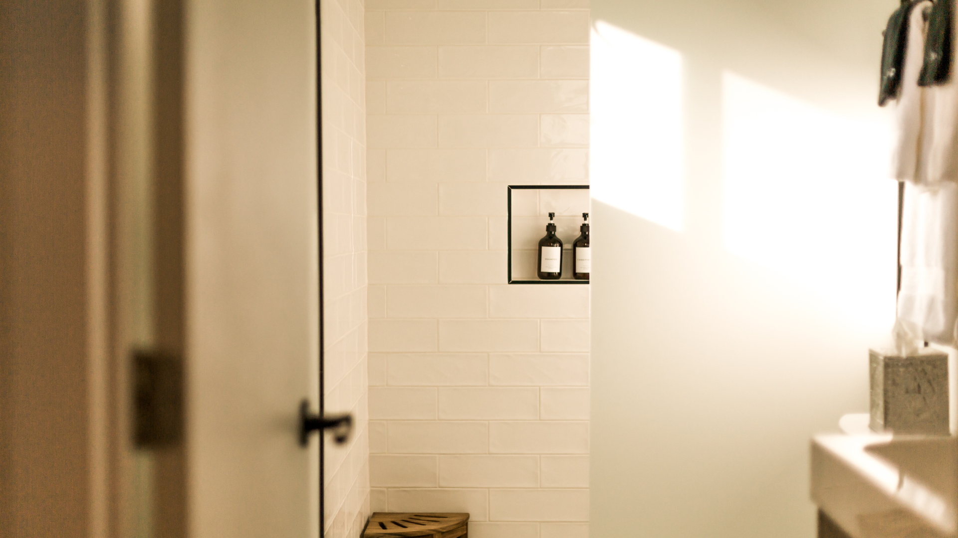View of a bathroom with beige tiled wall, a recessed shelf with two black bottles, and a wooden bench at the bottom corners, partially visible, with sunlight illuminating the space.