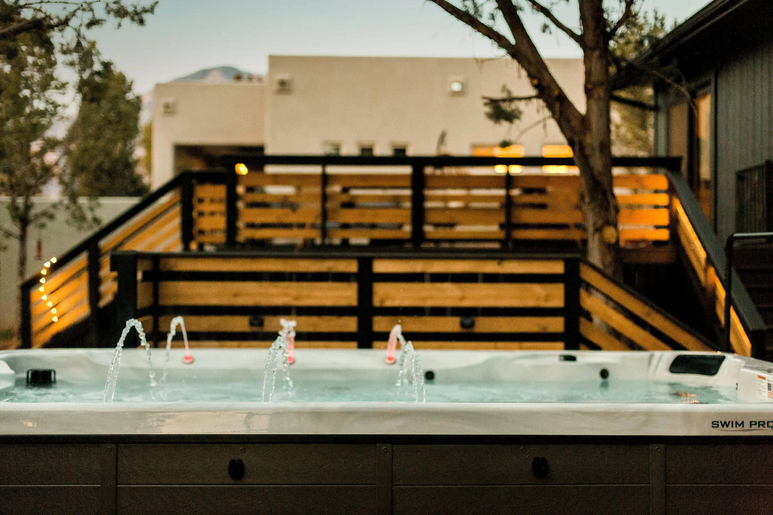 Hot tub with water jets on a patio, with a wooden deck and staircase, trees, and buildings in the background during dusk.