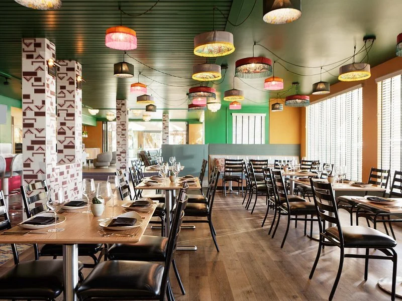 Empty restaurant dining area with wooden tables, black chairs, colorful hanging lamps, and large windows with blinds