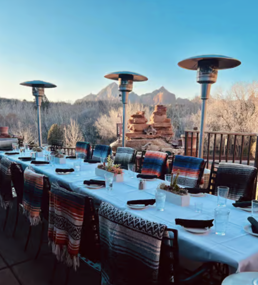 Outdoor dining table set with black chairs, black napkins, glasses, and small potted plants, overlooking a mountain landscape with heaters and desert rock formations.