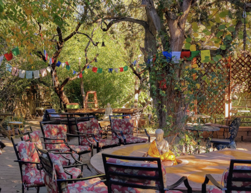 Outdoor patio with floral-patterned chairs, a Buddha statue, and colorful paper banners hanging from trees in a garden setting.