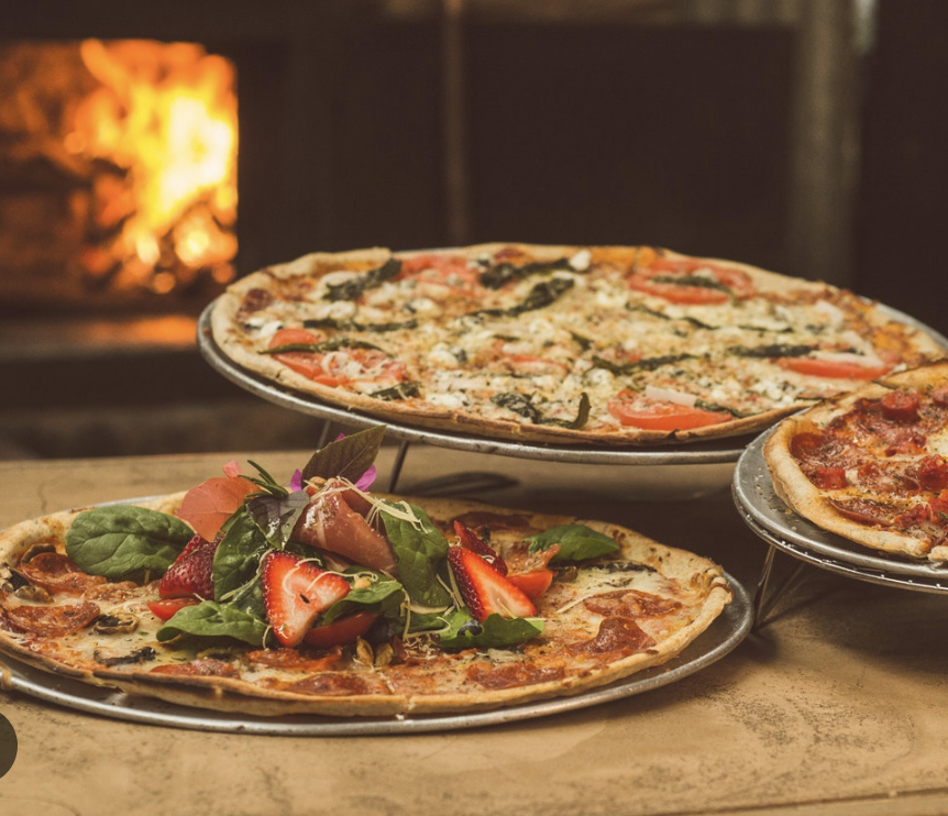 Three pizzas on display, one topped with fresh strawberries, greens, and prosciutto, another with pepperoni and cheese, and a third with spinach, tomato, and cheese, with a fireplace in the background.