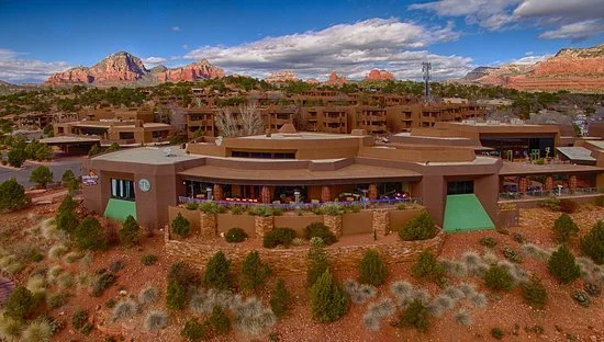 A desert landscape with multiple modern buildings and red rock formations in the background under a partly cloudy sky.